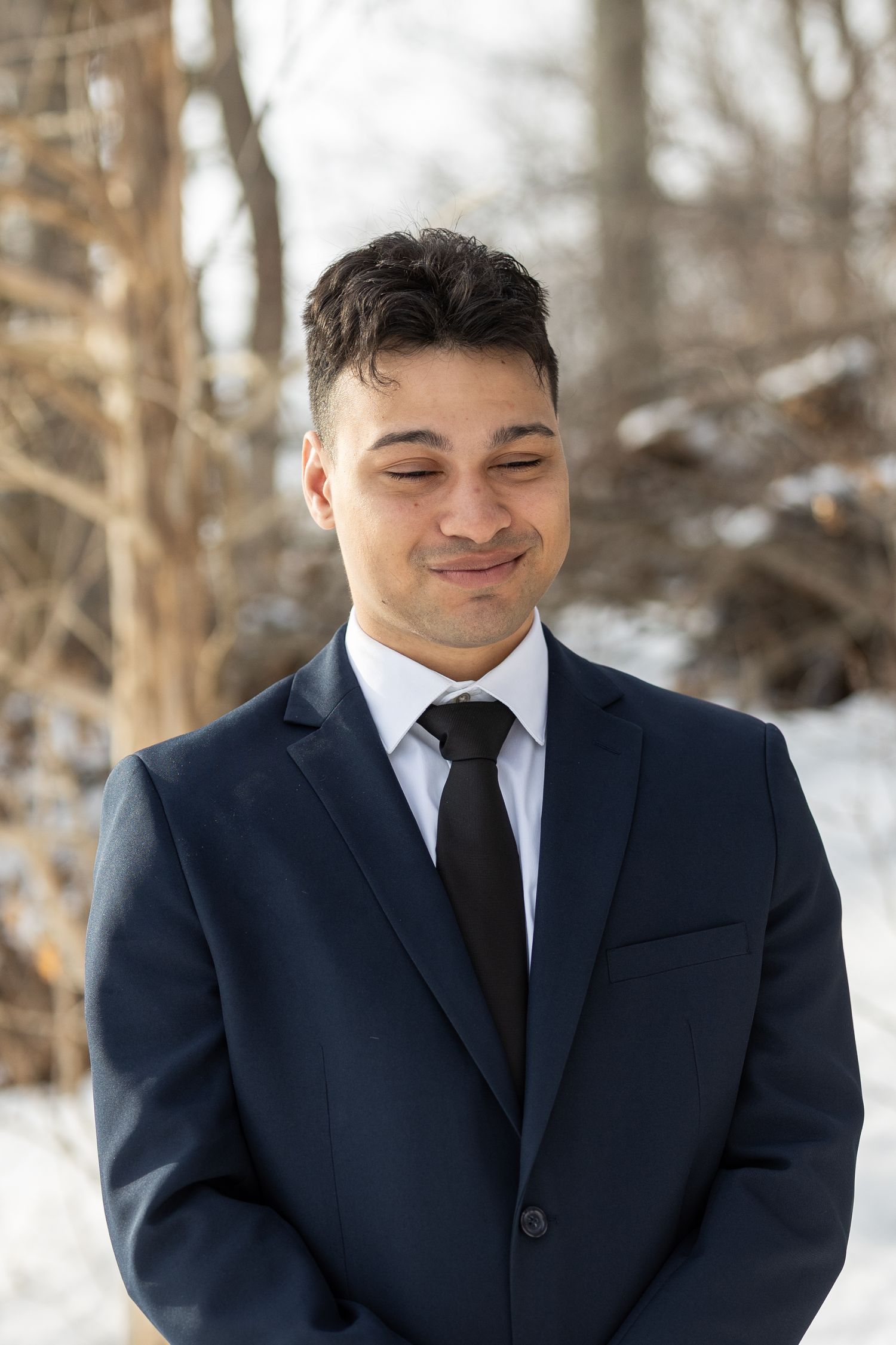 Young man in navy suit and black tie smiling outdoors in winter setting with snow and bare trees.
