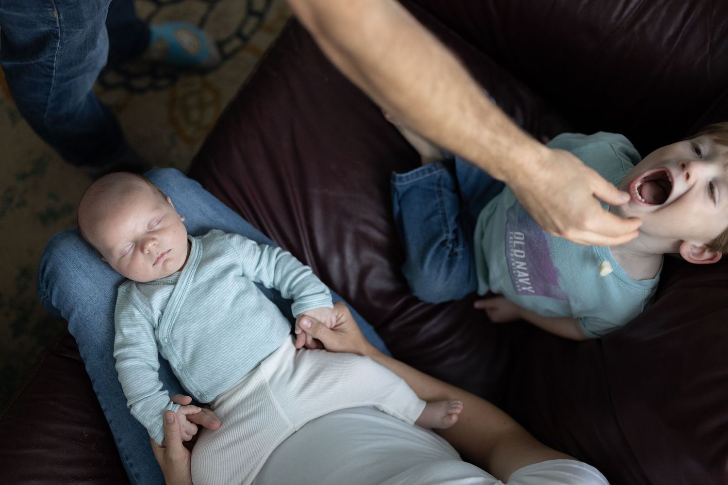 A baby wearing a light blue onesie sleeps peacefully on a caregiver's lap while being gently cradled. Father is feeding his other son.