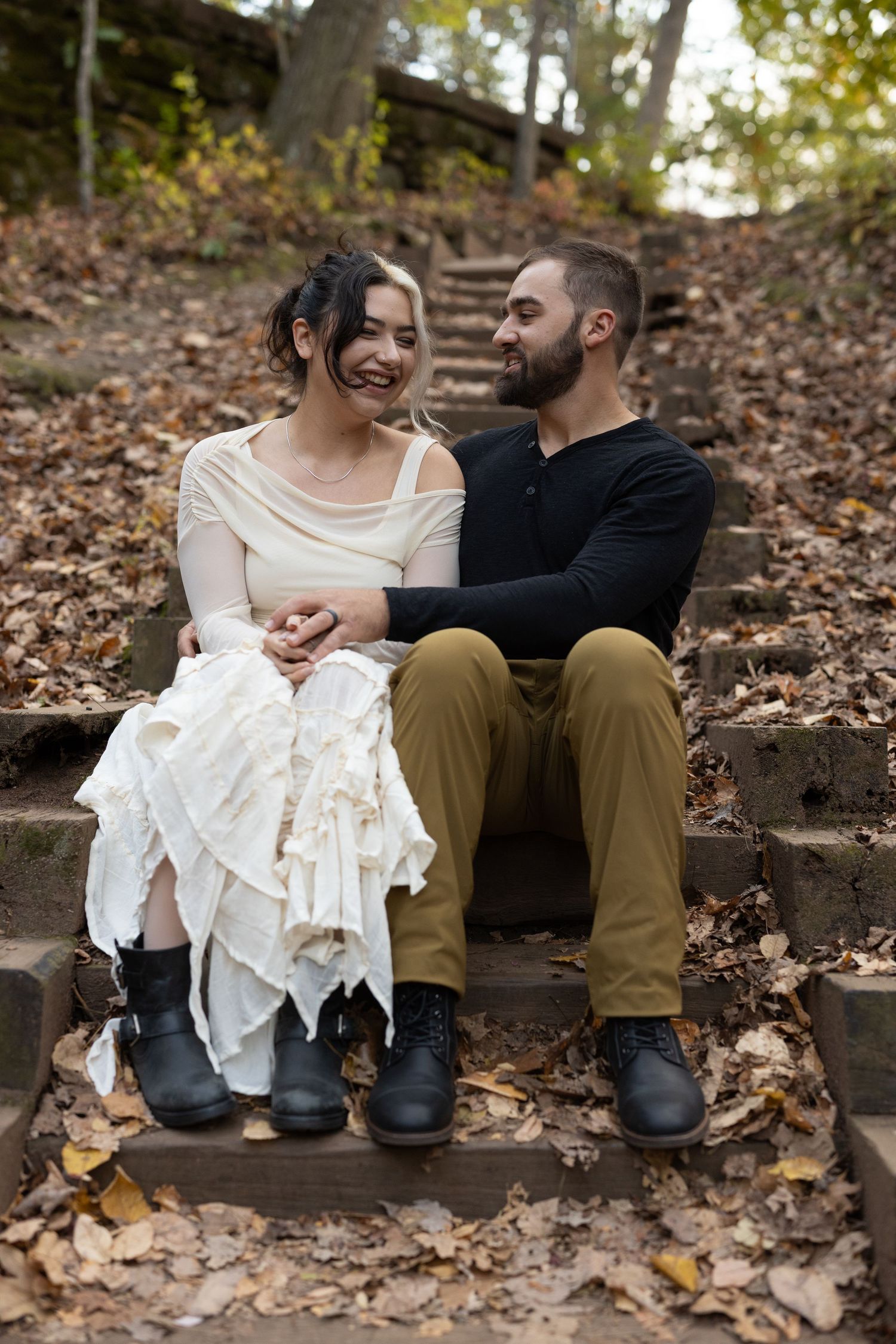 Couple sitting on autumn leaf-covered stone steps, smiling and holding hands, woman in white dress, man in black top.