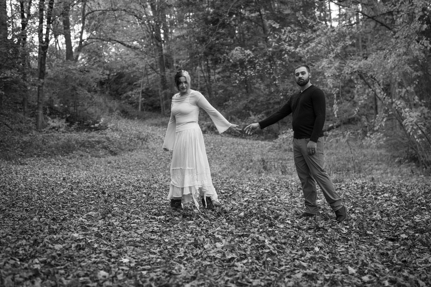 Black and white photo of a couple holding hands in a leaf-covered forest clearing, woman in white dress, man in dark sweater.