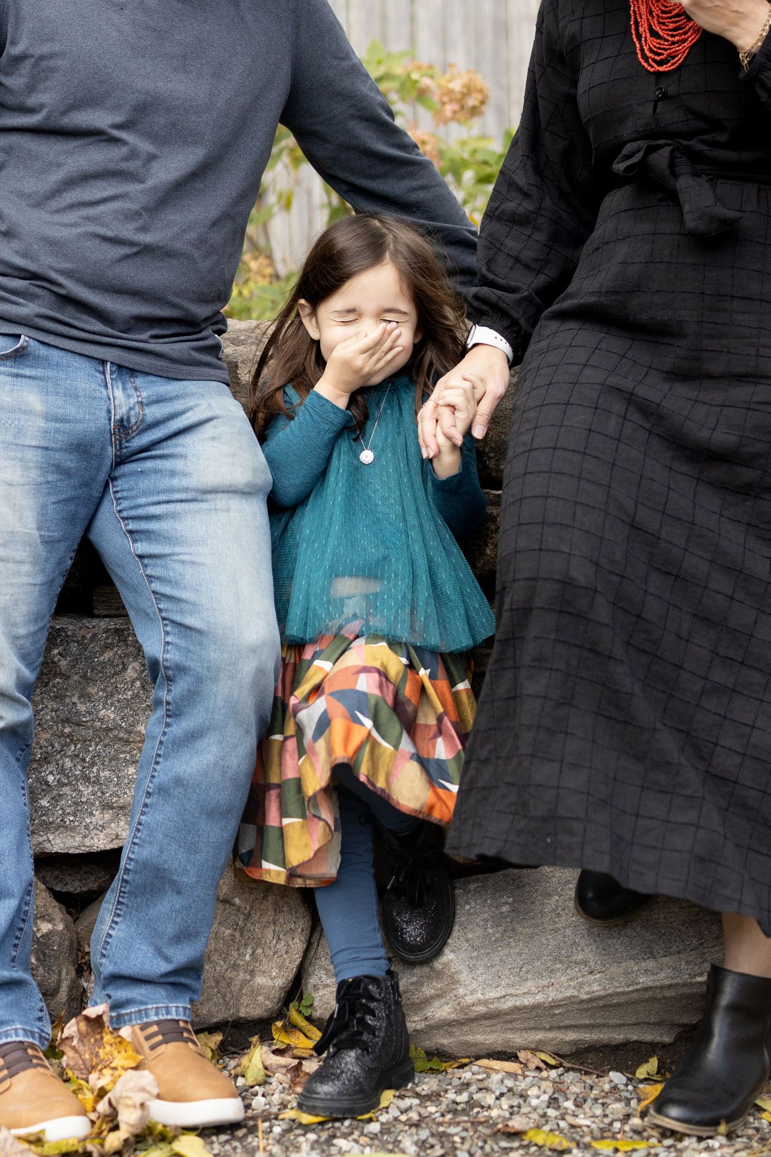 A child mid laughter as she covers her mouth. She is nestled between her mom and dads leg.