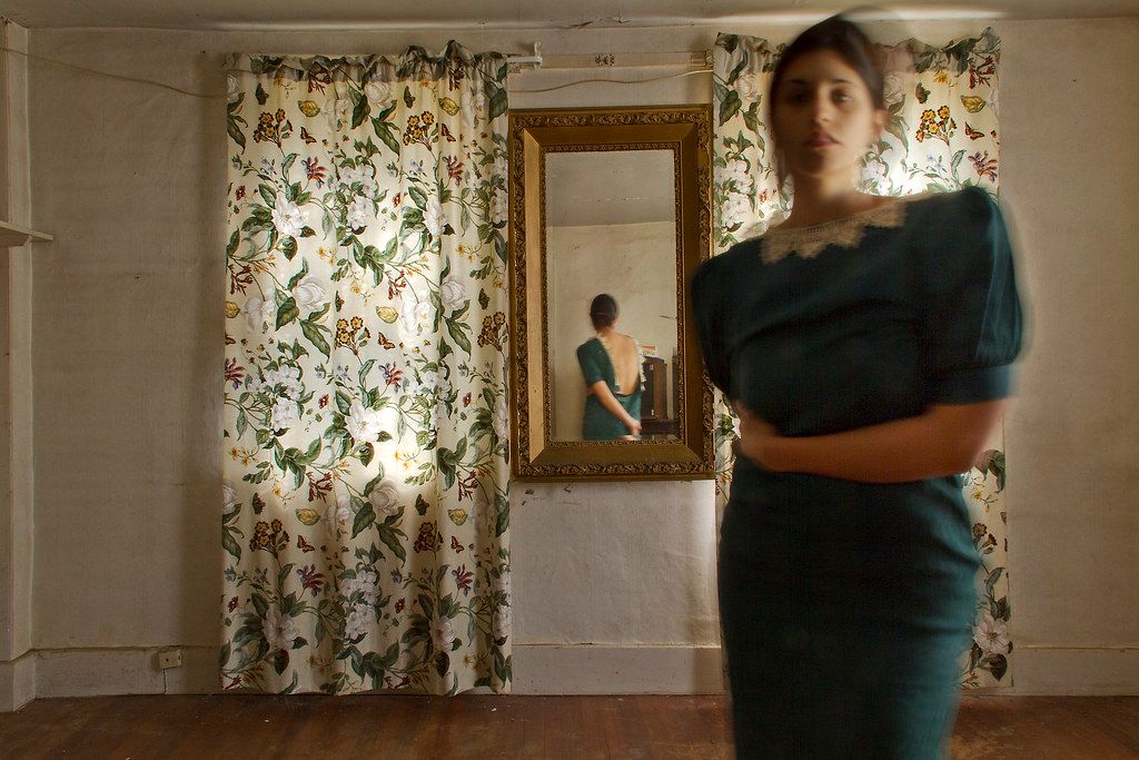Interior room with floral curtains and mirror reflecting a figure while another stands in teal dress.