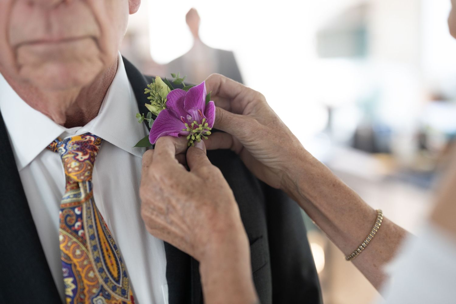 A person adjusts a colorful floral boutonniere on another person's formal suit lapel.