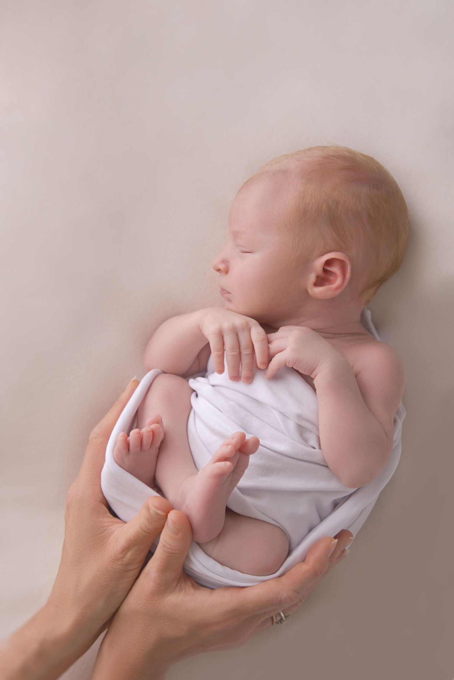 Newborn baby wrapped in a neutral swaddle and held by his mother's hands in a mobile studio setup in a Hasland home.
