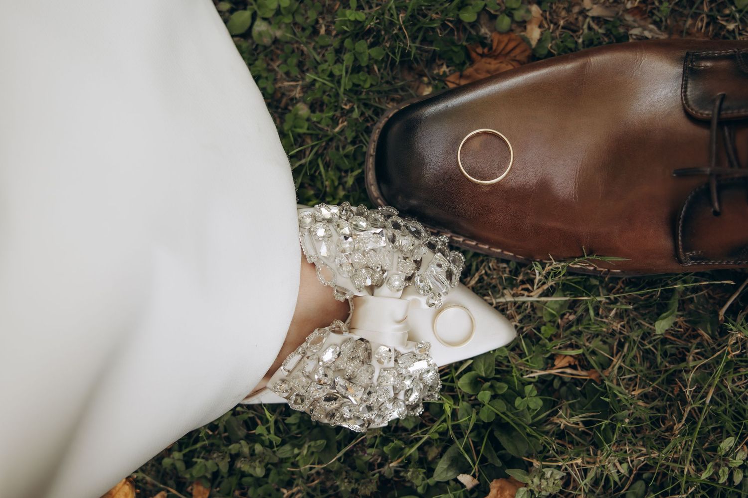 Wedding rings resting on brown leather dress shoe and bejeweled bridal heel on green grass.
