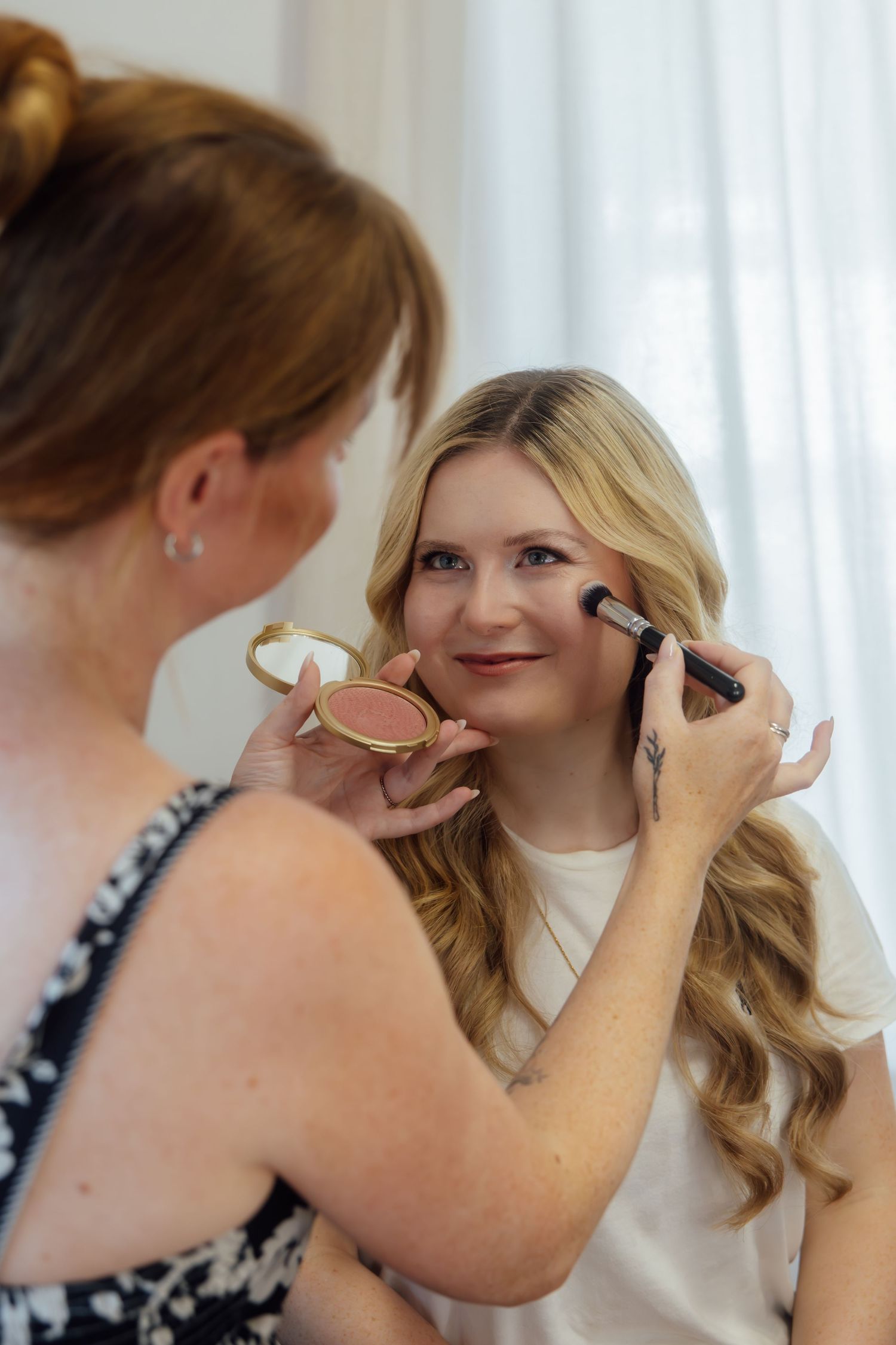 A makeup artist applies blush to a smiling blonde woman using a brush, holding a compact powder in a bright room.