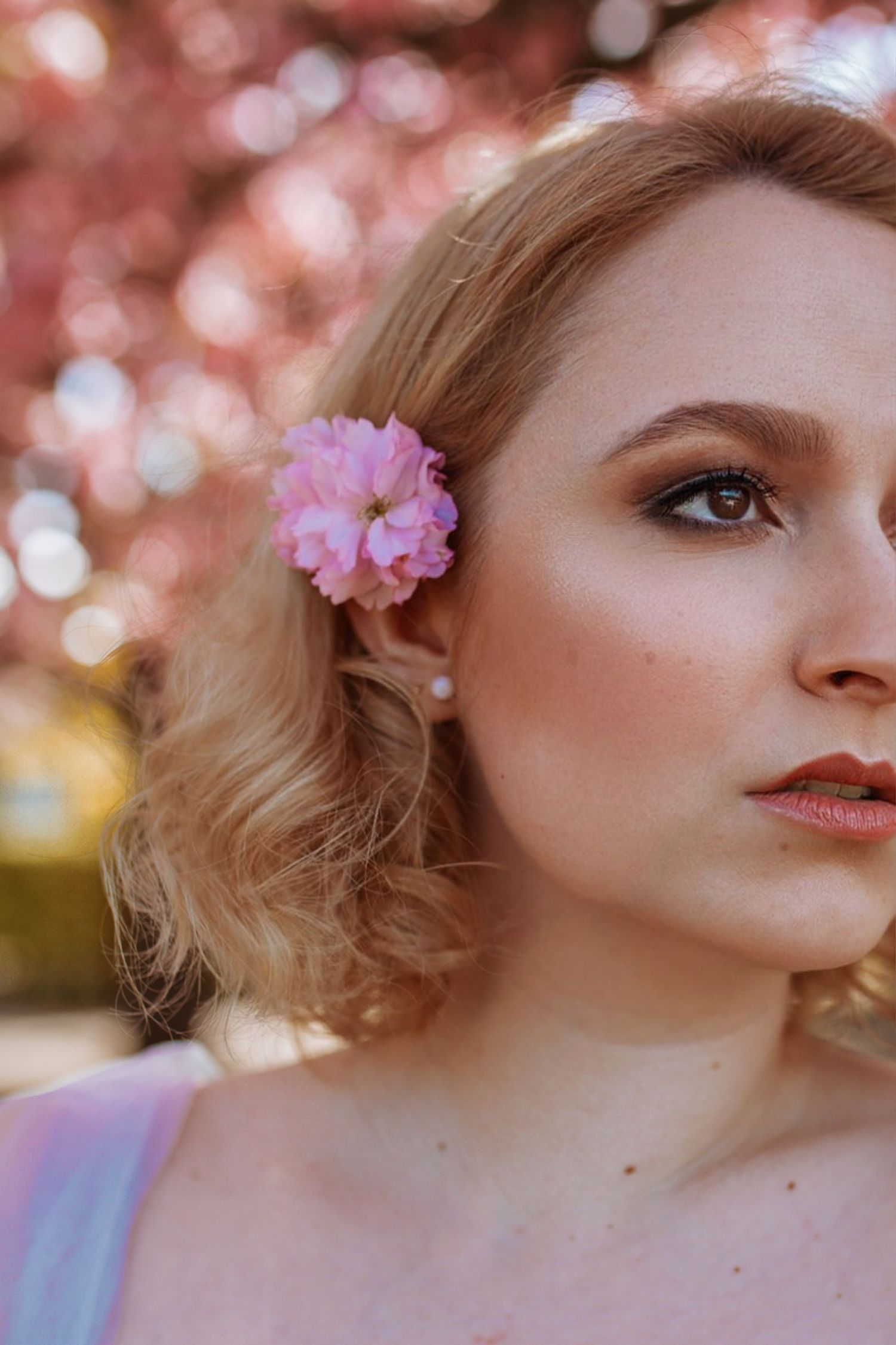 Close-up portrait of a blonde woman with a pink flower in her hair against a blossoming background.