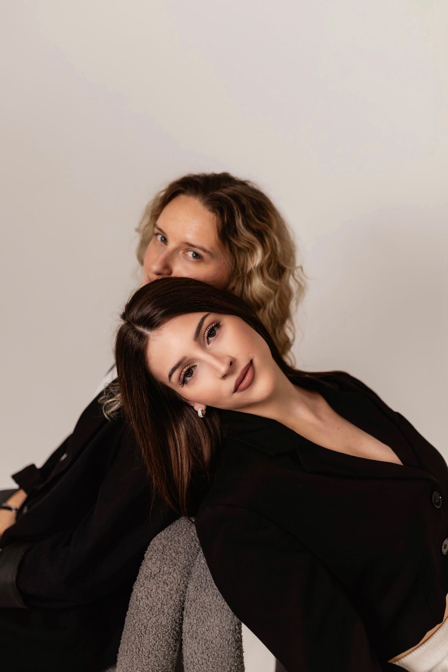 Two women in black outfits pose together against white background, one leaning on the other's shoulder.