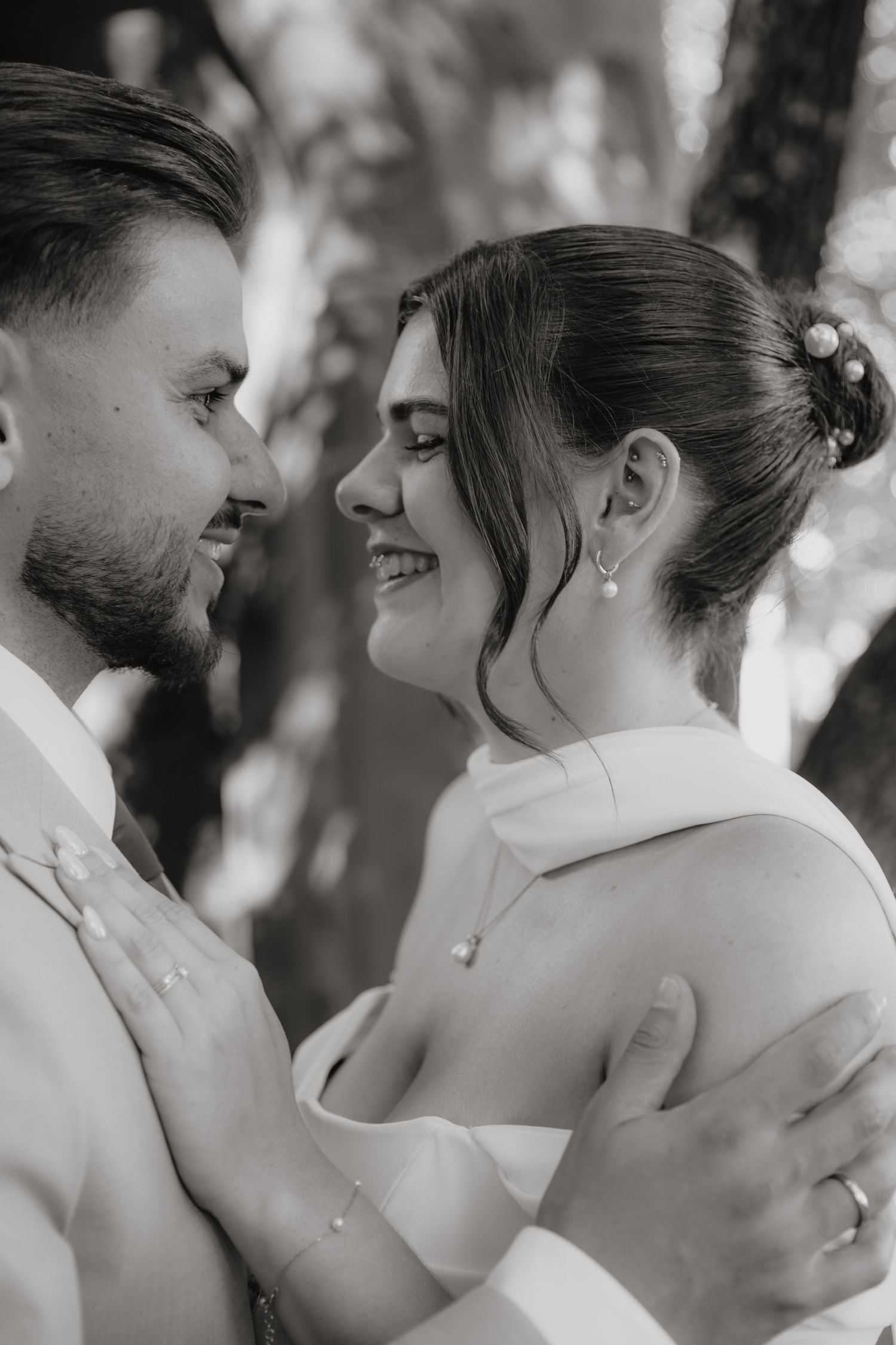 Black and white photo of smiling bride and groom embracing face to face outdoors among trees.