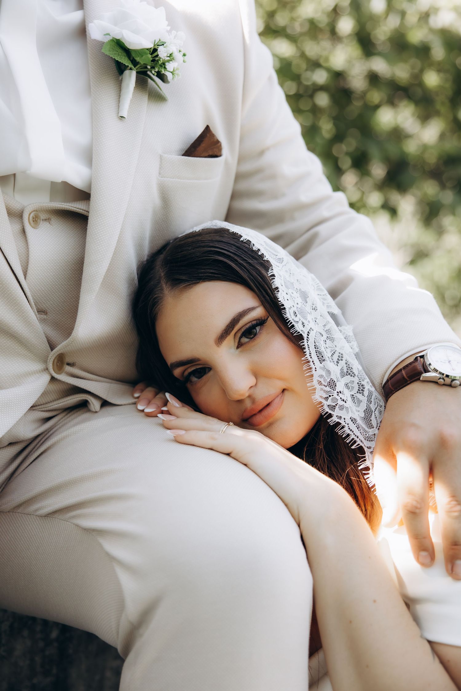 Bride in lace veil rests head on groom's knee in beige suit with boutonniere during outdoor wedding photo.