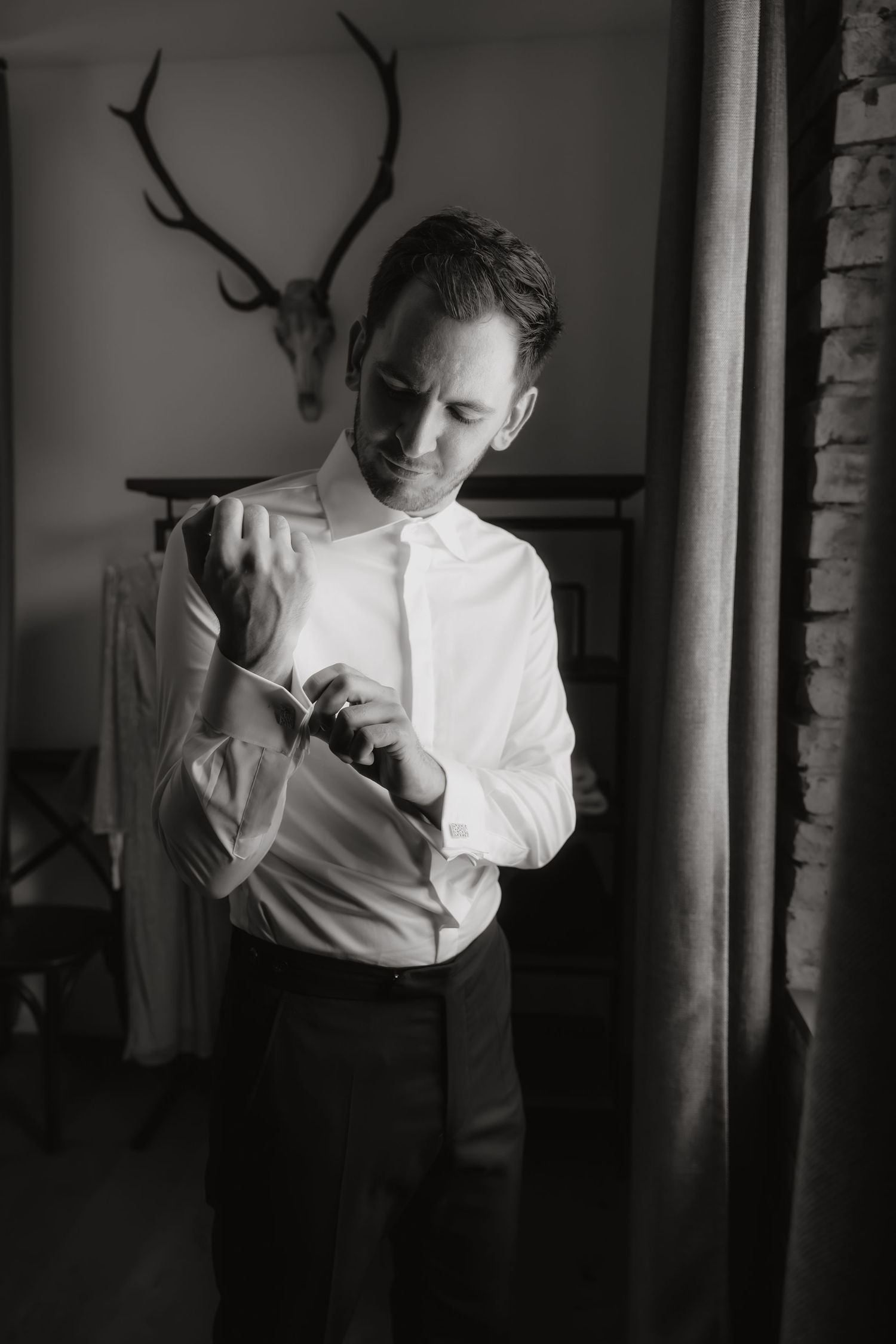 Groom in white dress shirt fastening cufflinks in a rustic room with antler decor in black and white photo.