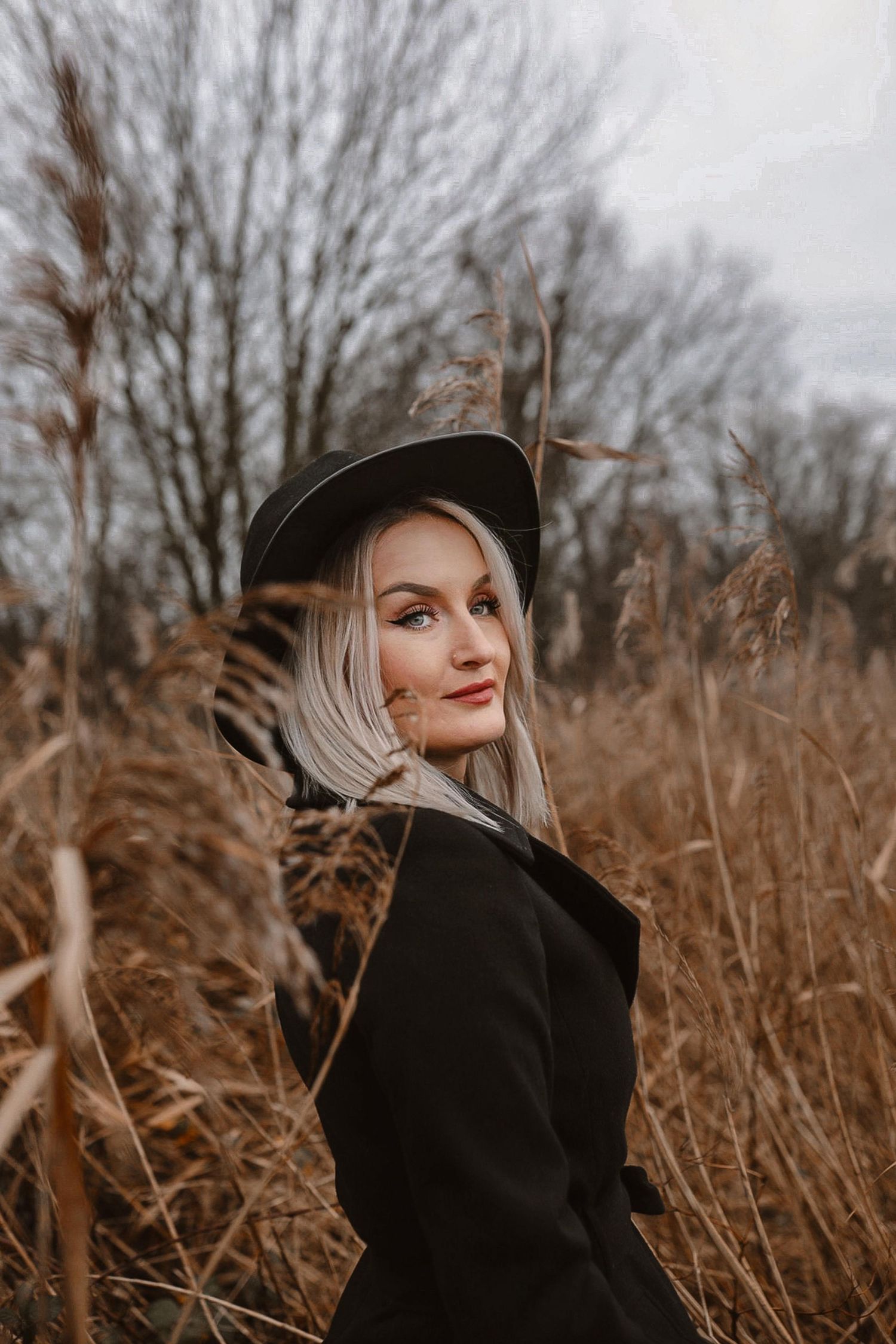 A blonde woman wearing a black hat and coat posing among tall dry grass in an autumn field.
