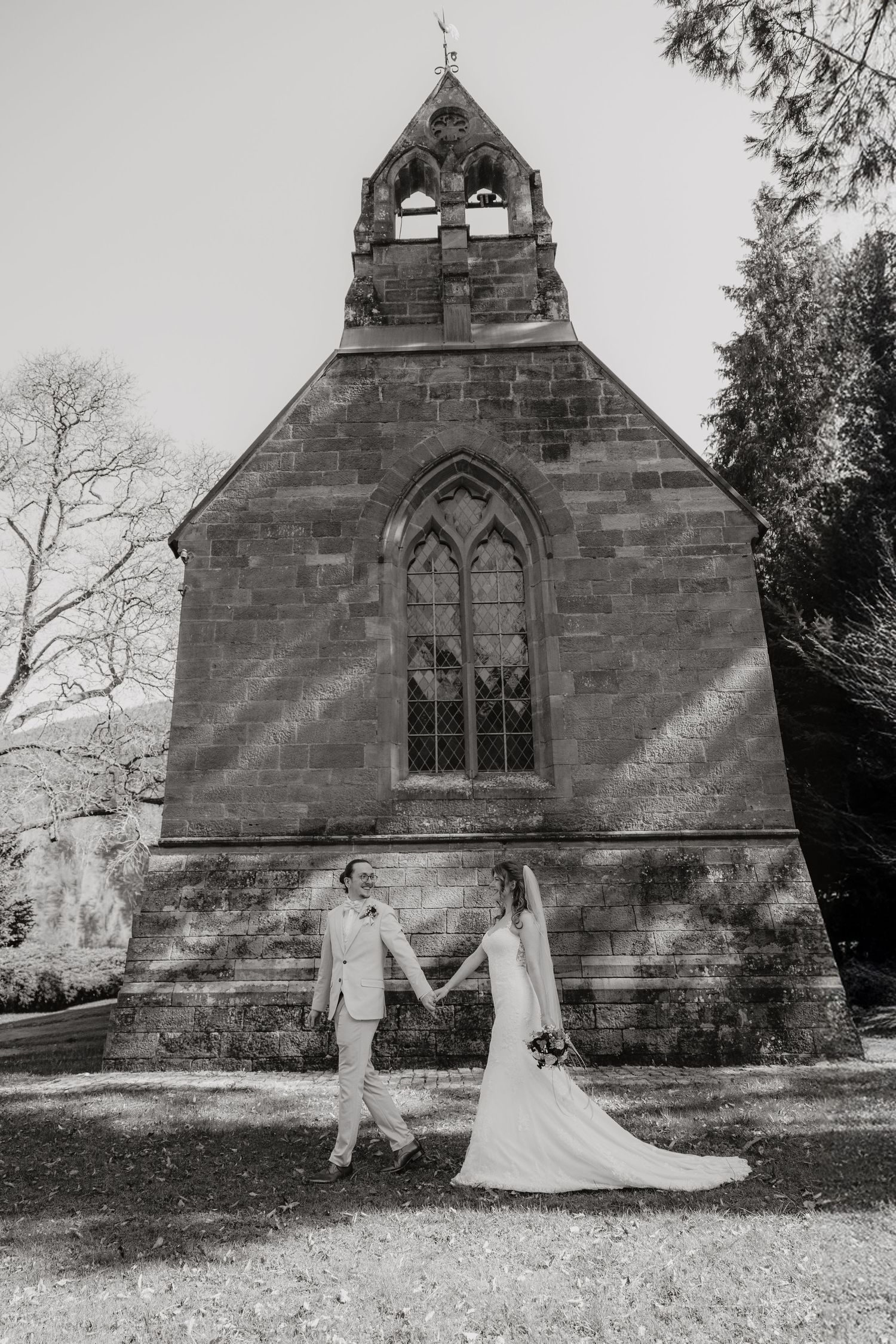 Bride and groom holding hands in black and white photo before stone chapel.