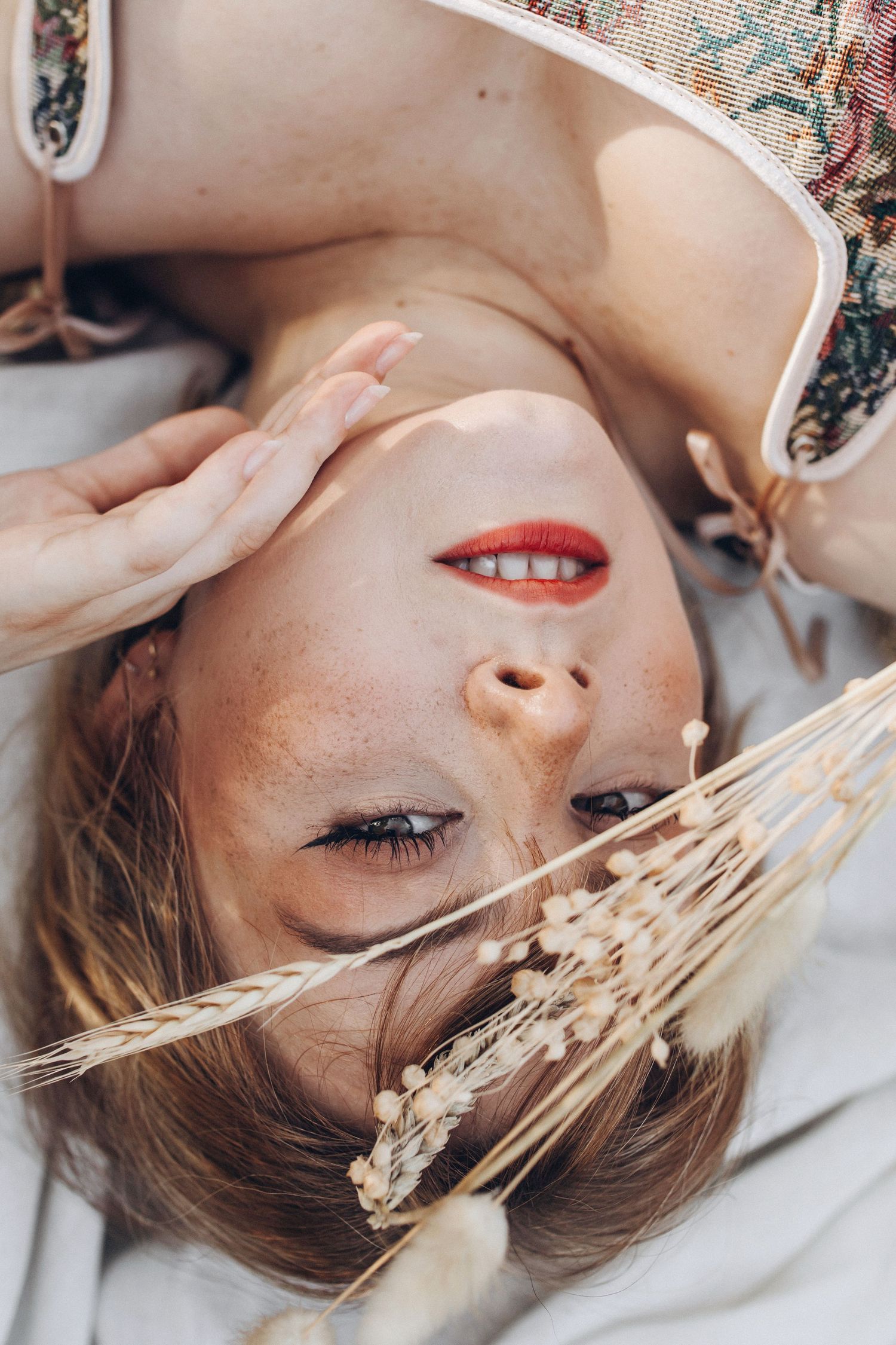 Close-up of freckled woman with red lips lying upside down in wheat field with dried flowers.
