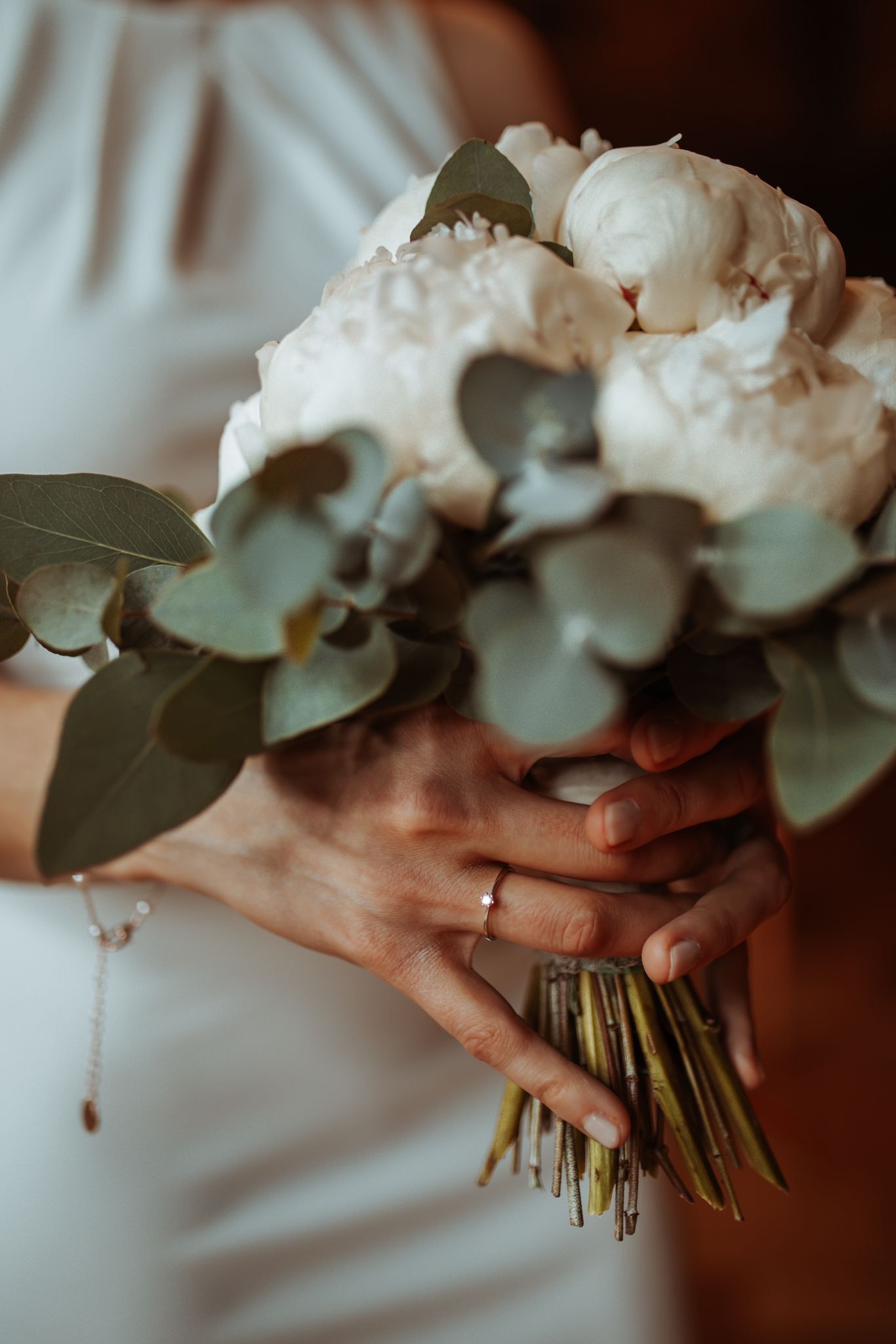 Bride holding elegant wedding bouquet of white peonies and eucalyptus, wearing a diamond ring on her finger.