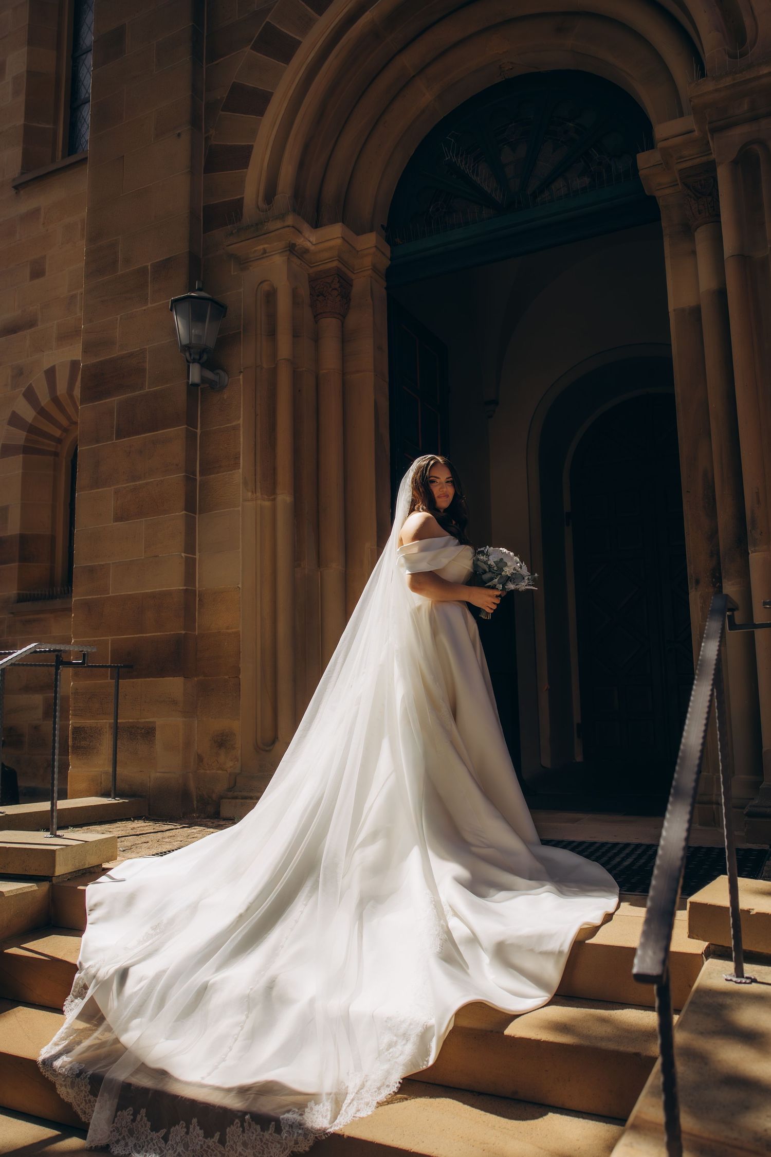 Bride in flowing white gown and veil stands on church steps, holding bouquet, with sunlit sandstone architecture.