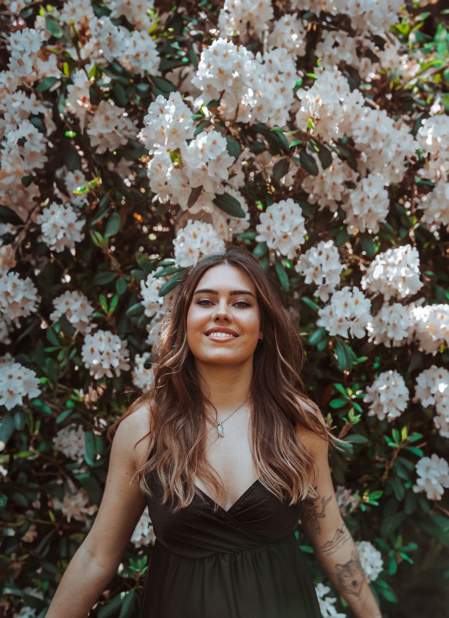 A smiling brunette woman with a tattoo stands before a wall of white blooming flowers.