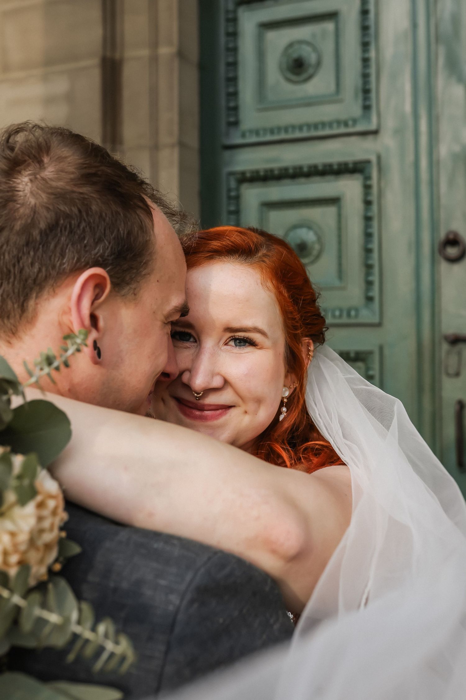 Radiant red-haired bride with veil smiling over grooms shoulder during wedding embrace near ornate door.