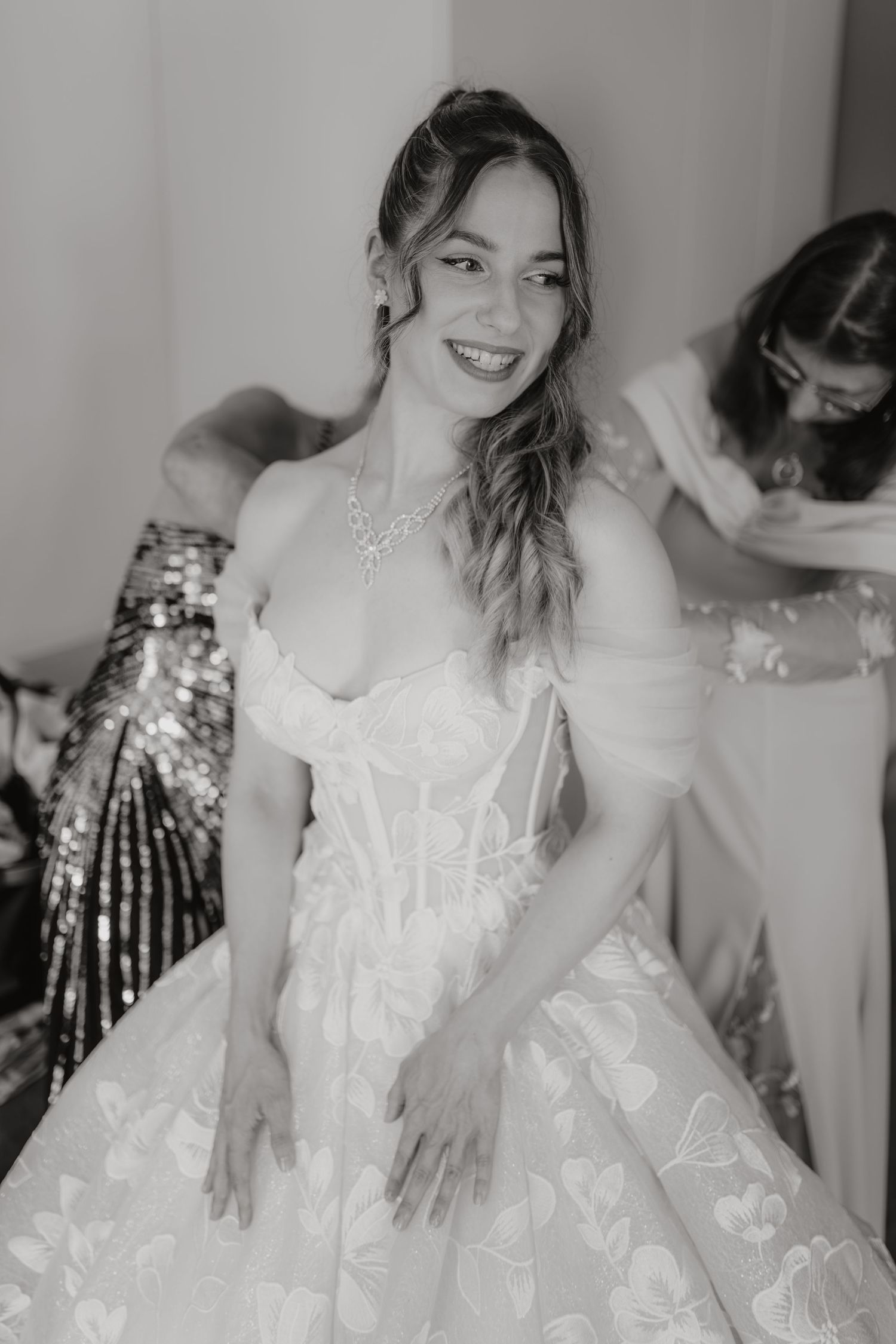 Smiling bride in a floral ball gown wedding dress being helped by bridesmaids, black and white photo.