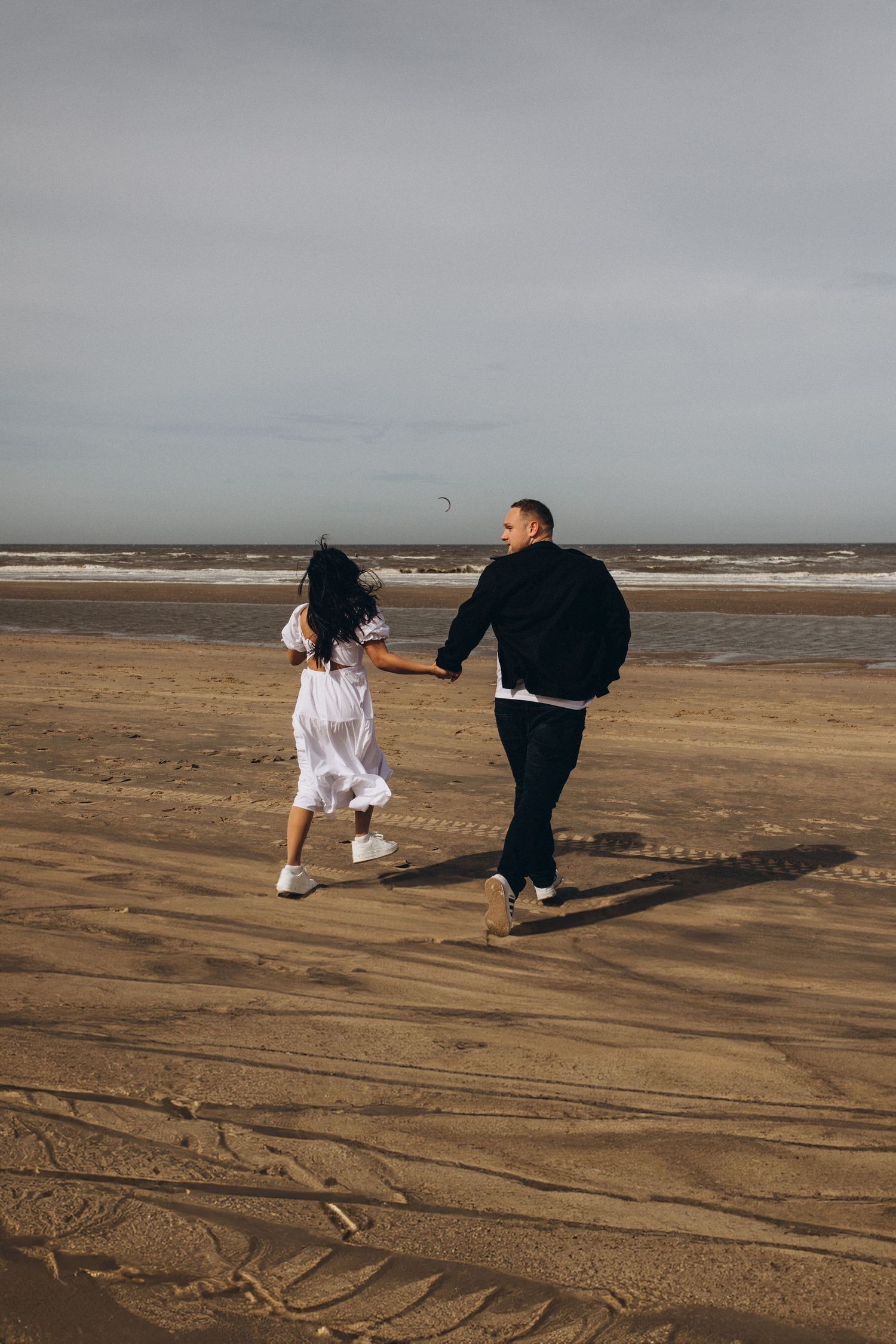 Couple holding hands running on a wide sandy beach under cloudy skies.