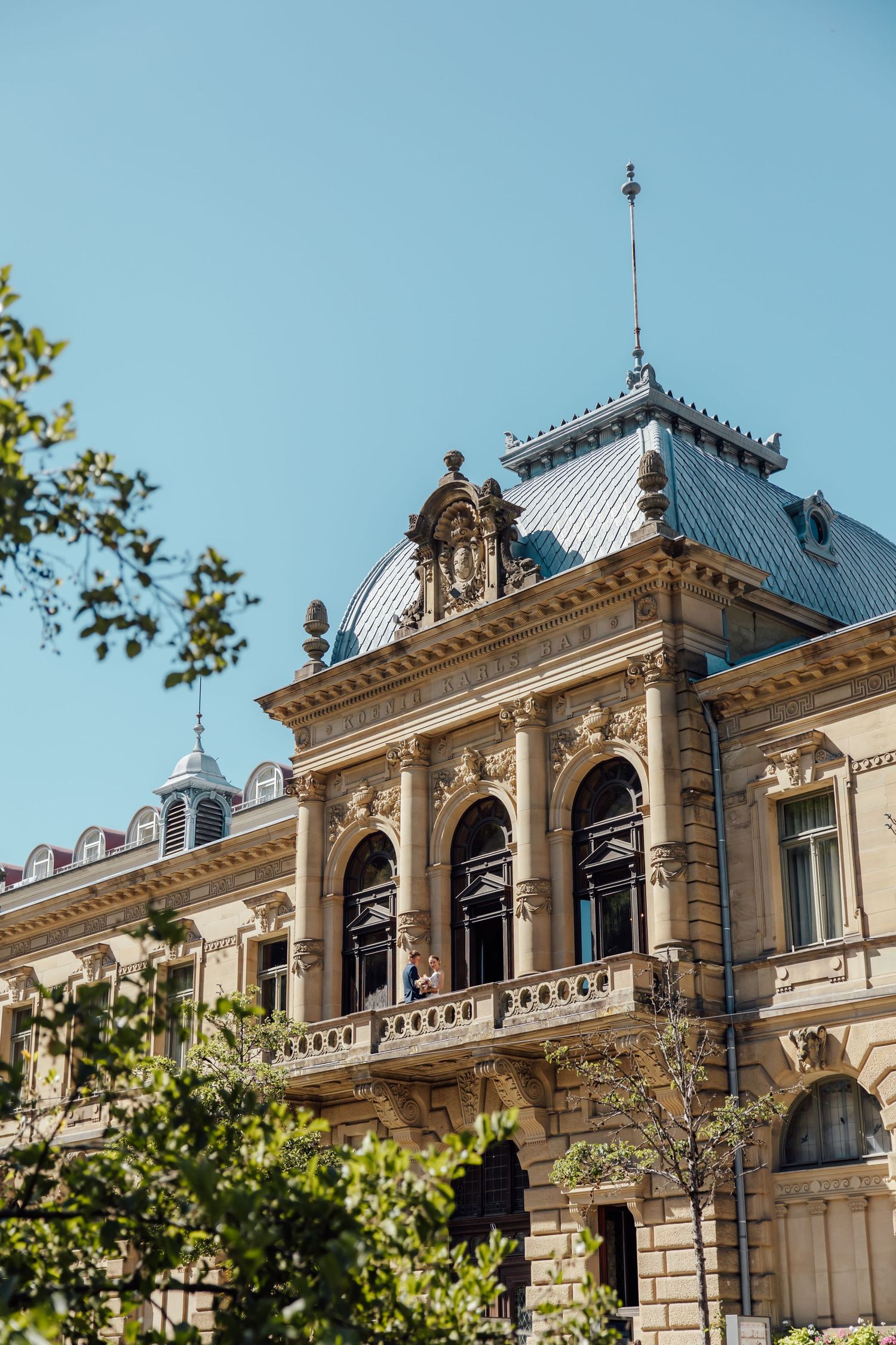 Ornate Beaux-Arts building facade with arched windows, decorative balcony, and mansard roof against clear blue sky.