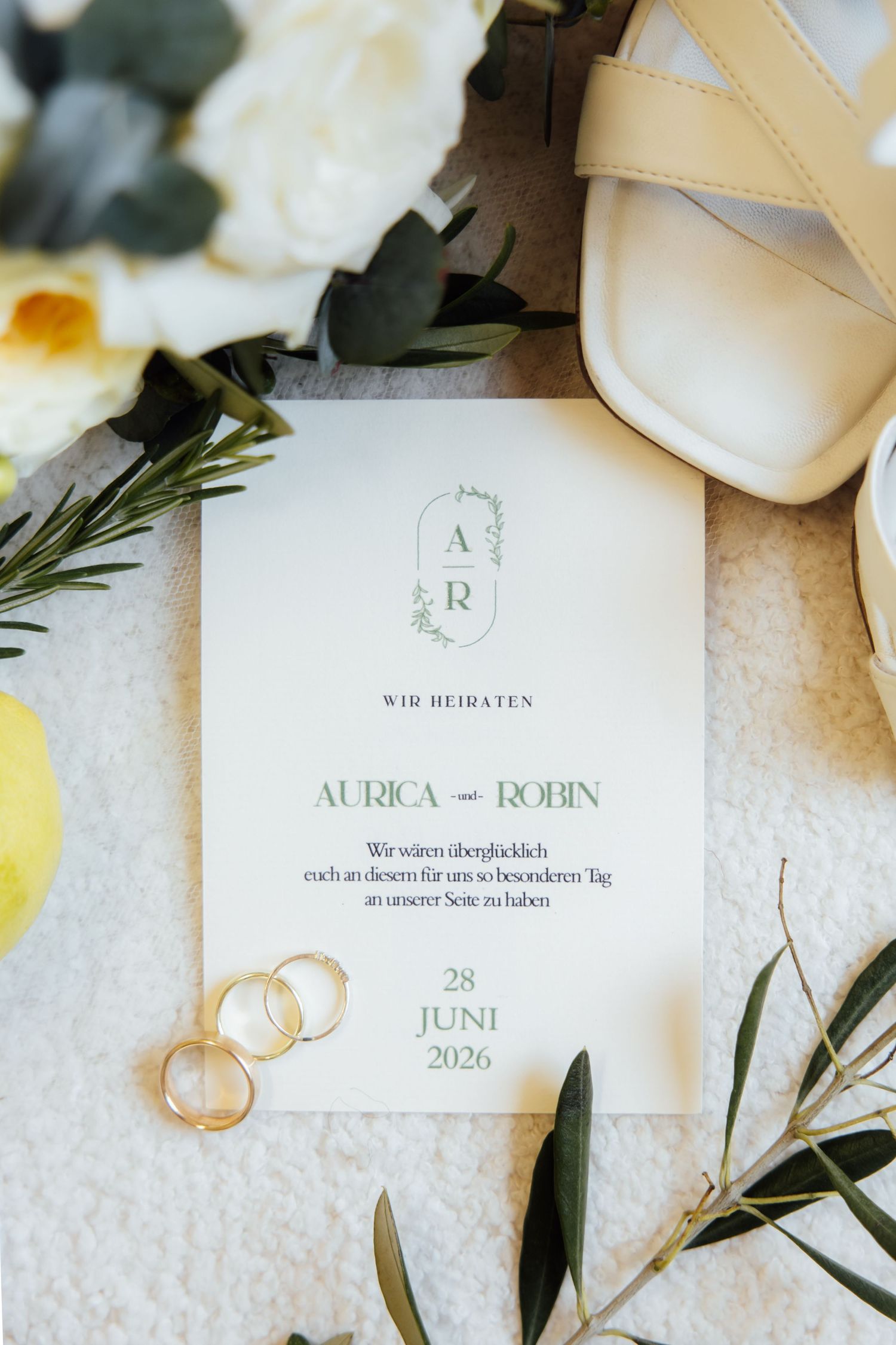 German wedding invitation for Aurica and Robin on 28 Juni 2026, surrounded by gold rings, white heels, and flowers.