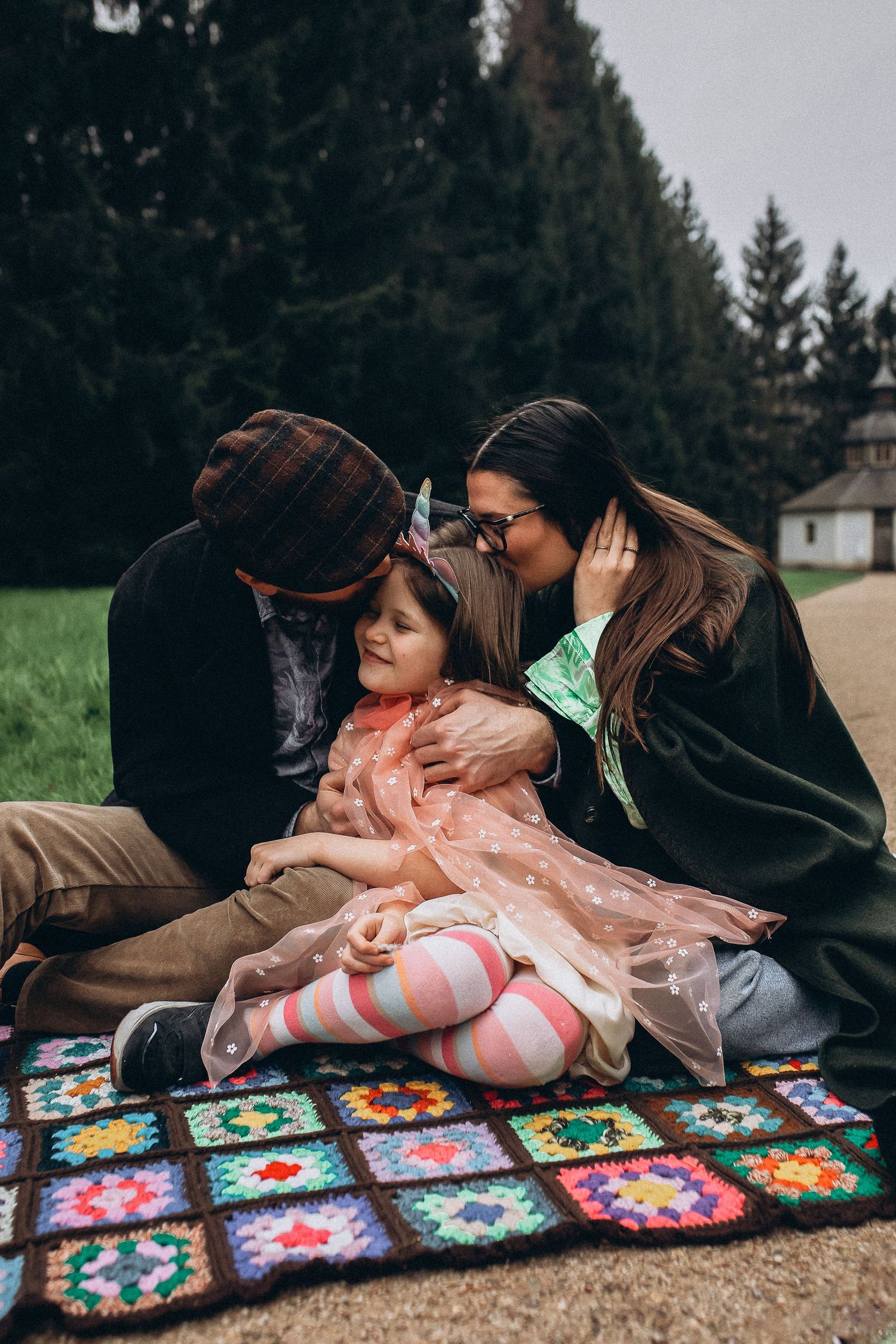 Family of three sharing kisses on a colorful blanket in the forest.