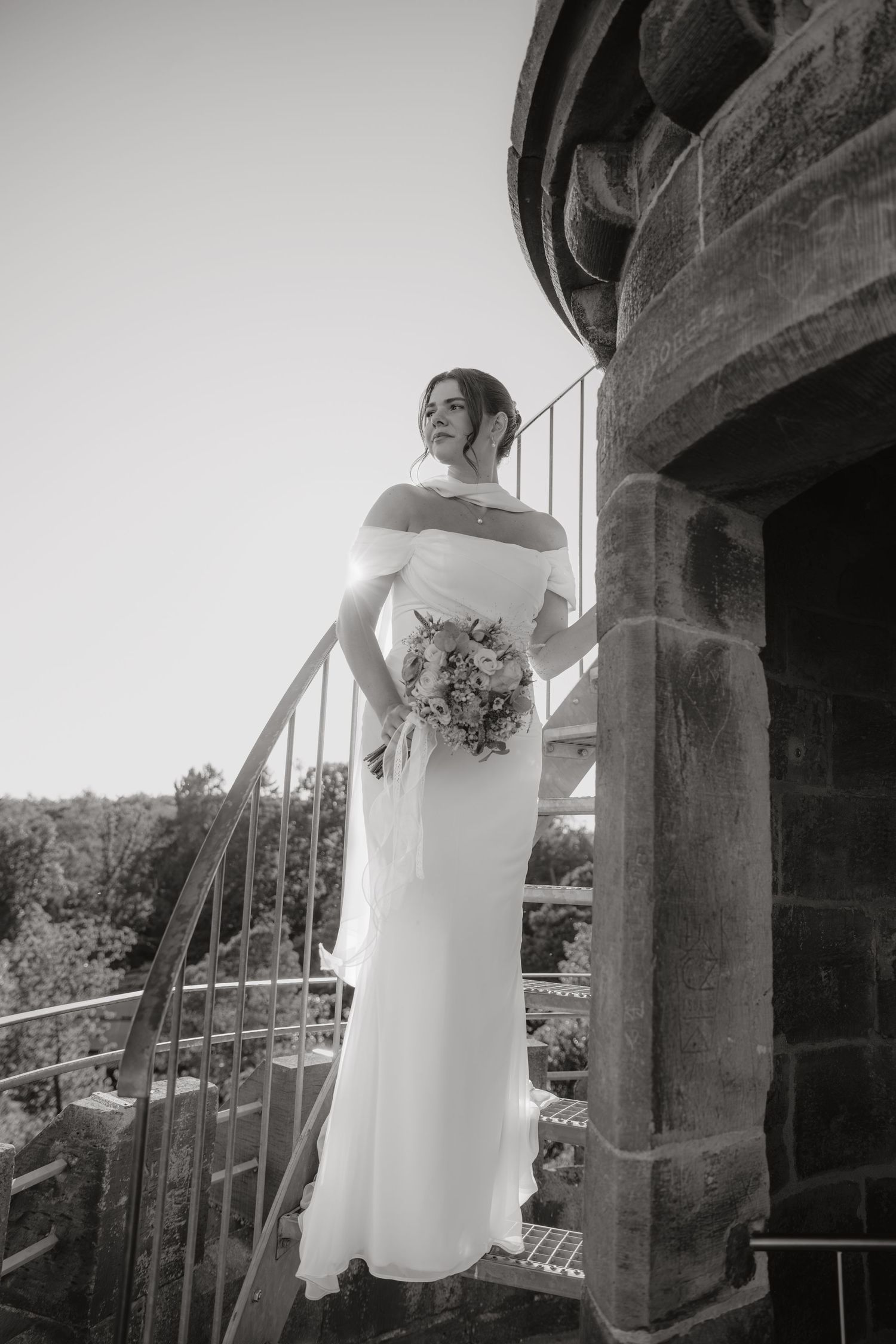 Elegant bride in off-shoulder gown holding bouquet on stone spiral staircase in black and white.