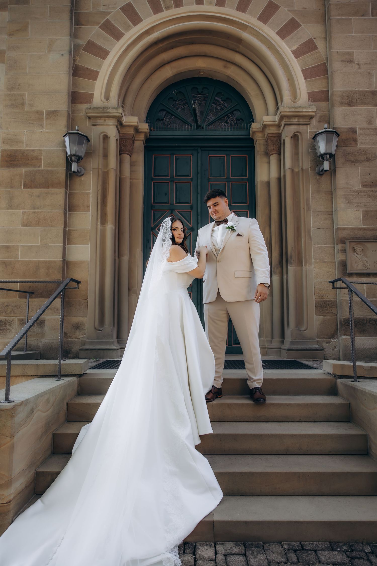 Bride in white gown and groom in beige suit pose on church steps before ornate green door.