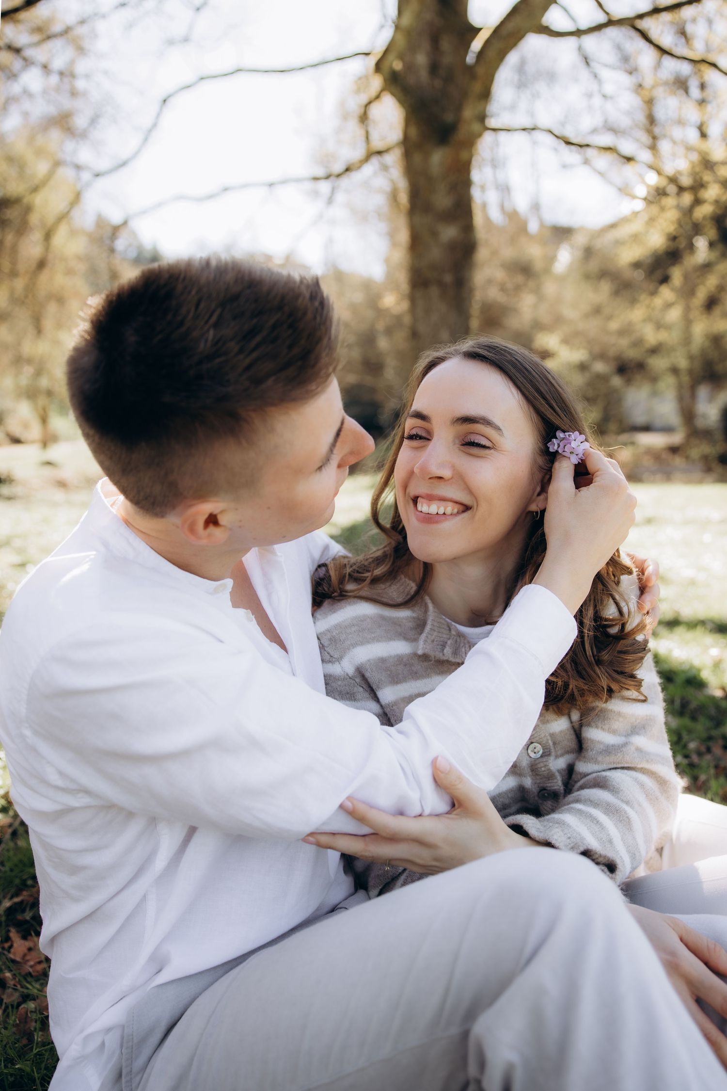 A couple sitting outdoors in a park, smiling as a man tucks a small purple flower behind a woman's ear.
