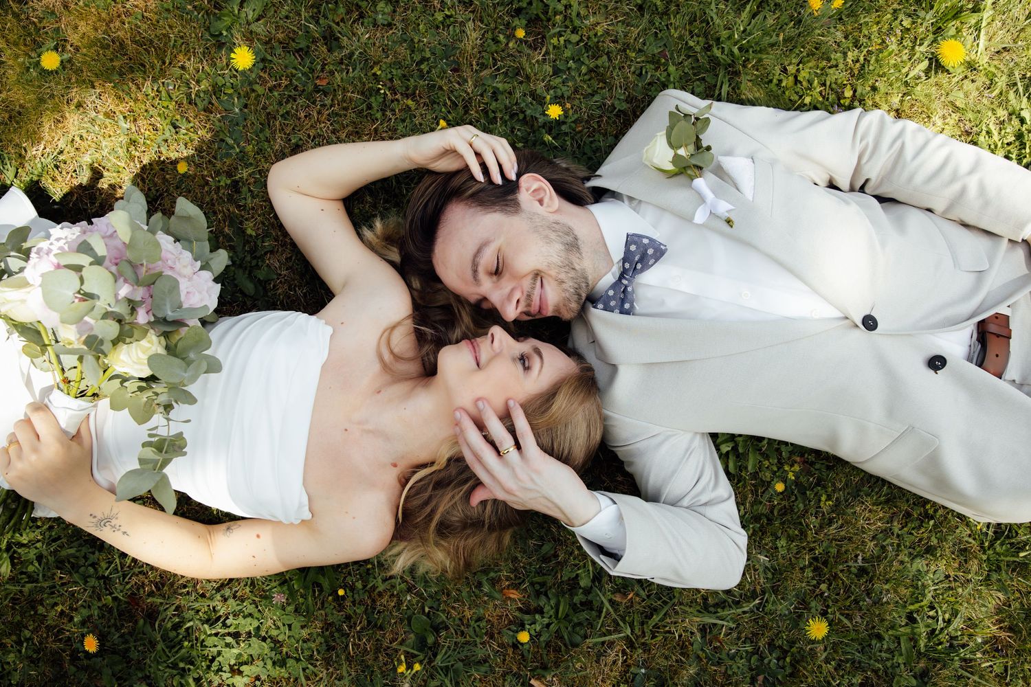 Bride and groom lying on green grass, smiling at each other, bride holding white bouquet, groom in light suit with bow tie.