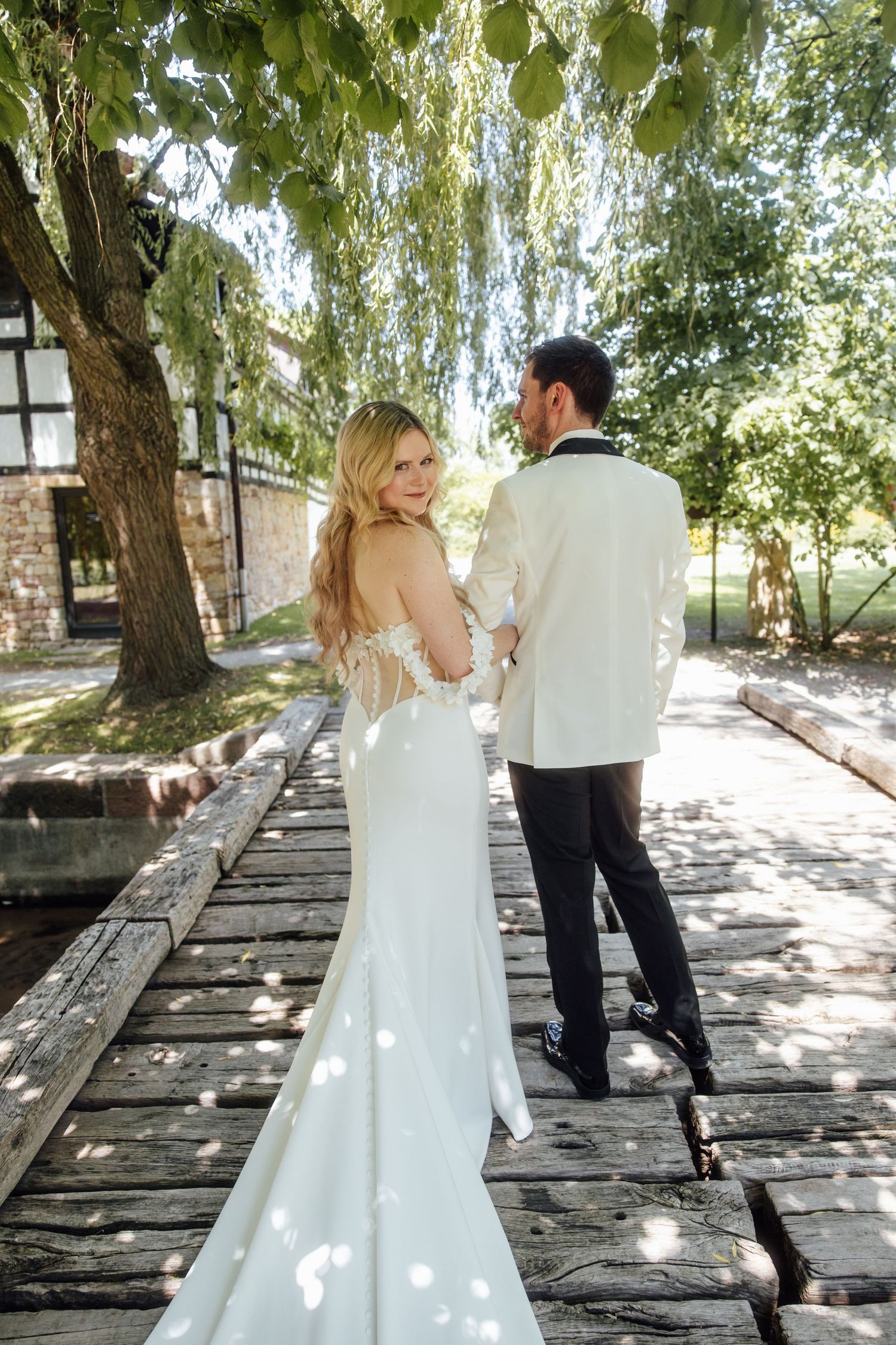 Bride in backless white gown and groom in white tuxedo walking on wooden bridge surrounded by lush green trees.