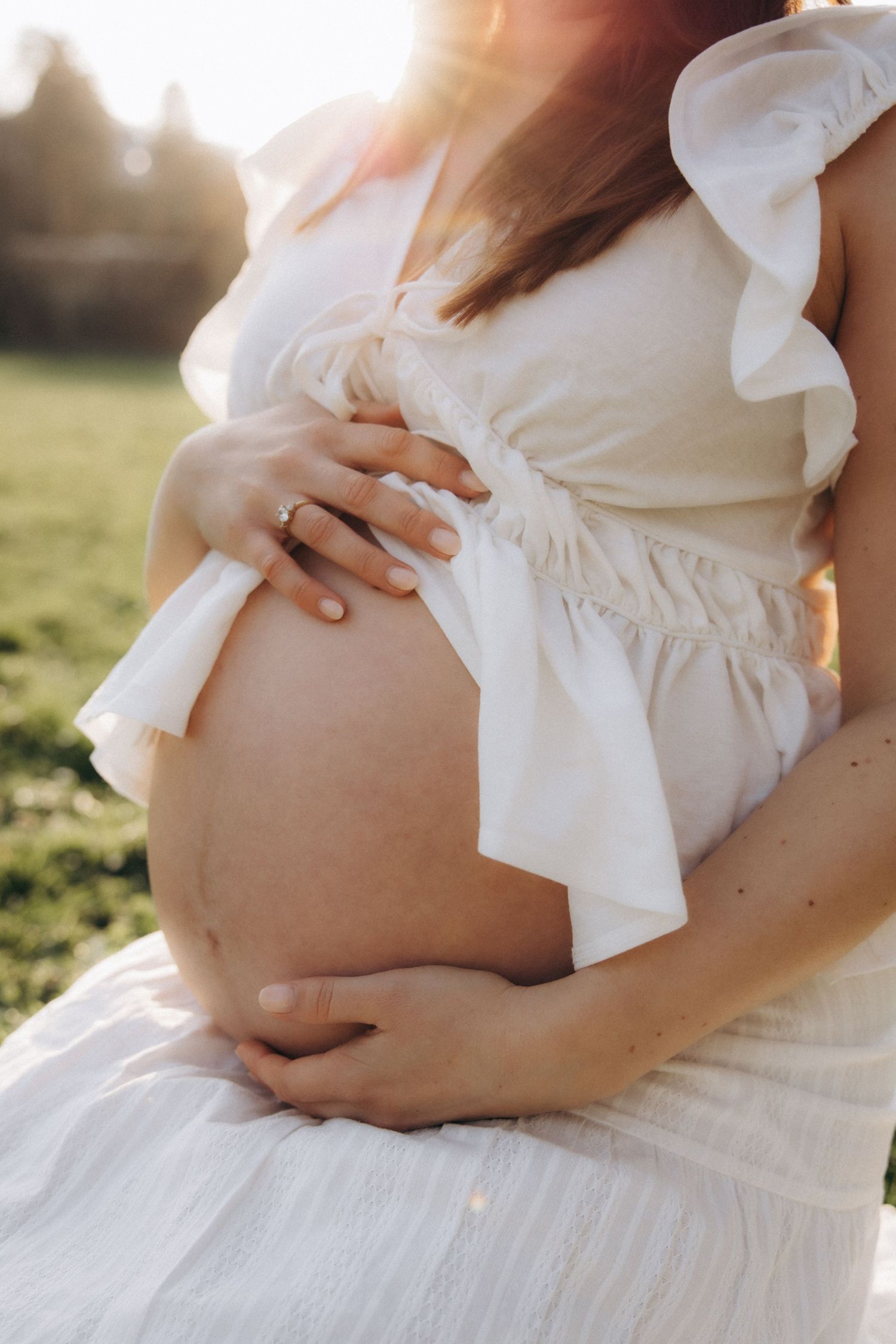 Close-up of pregnant woman in white ruffled dress cradling bare baby bump outdoors.