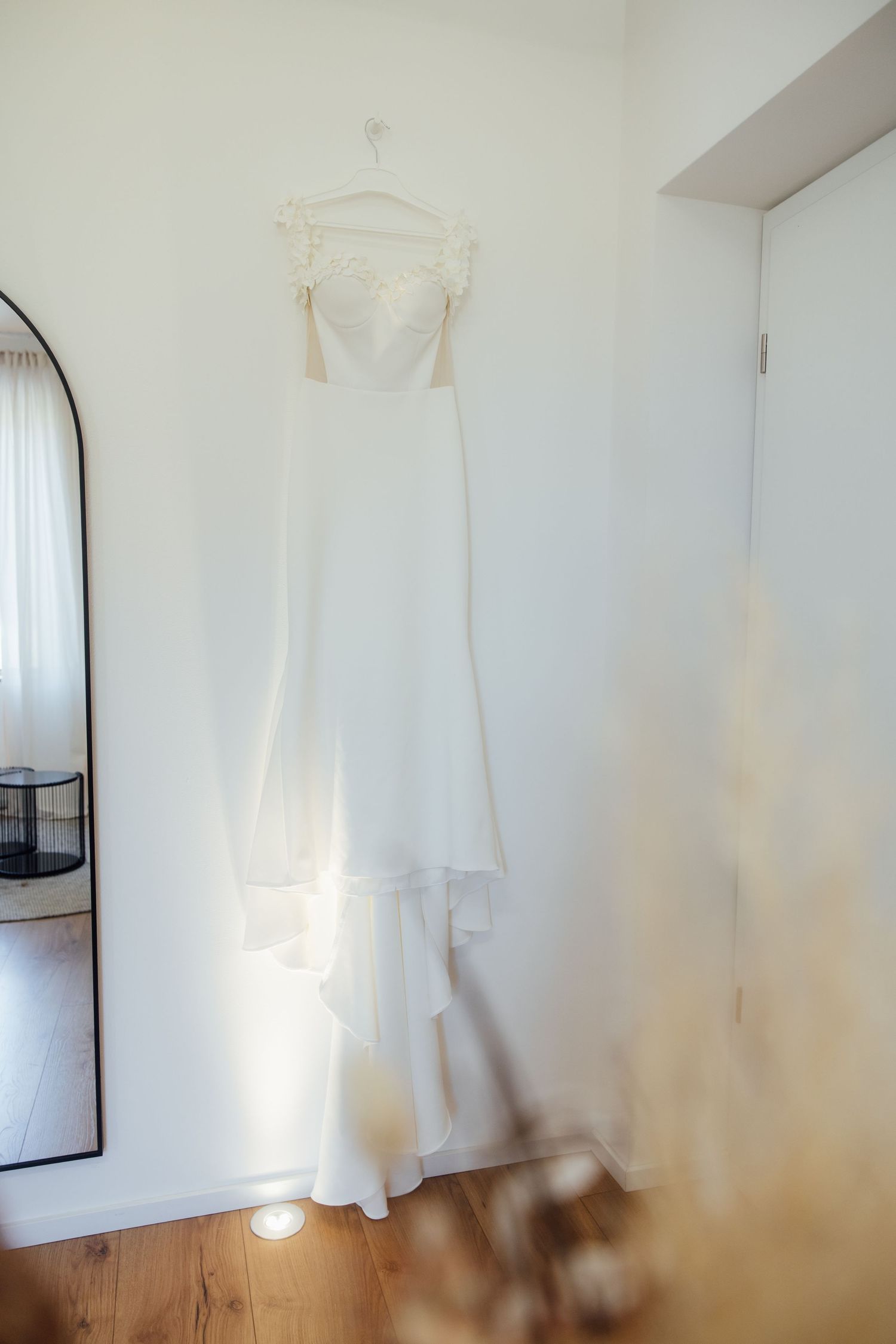 Elegant white wedding dress with lace bodice hanging on a wall hanger in a bright, minimalist bridal suite.