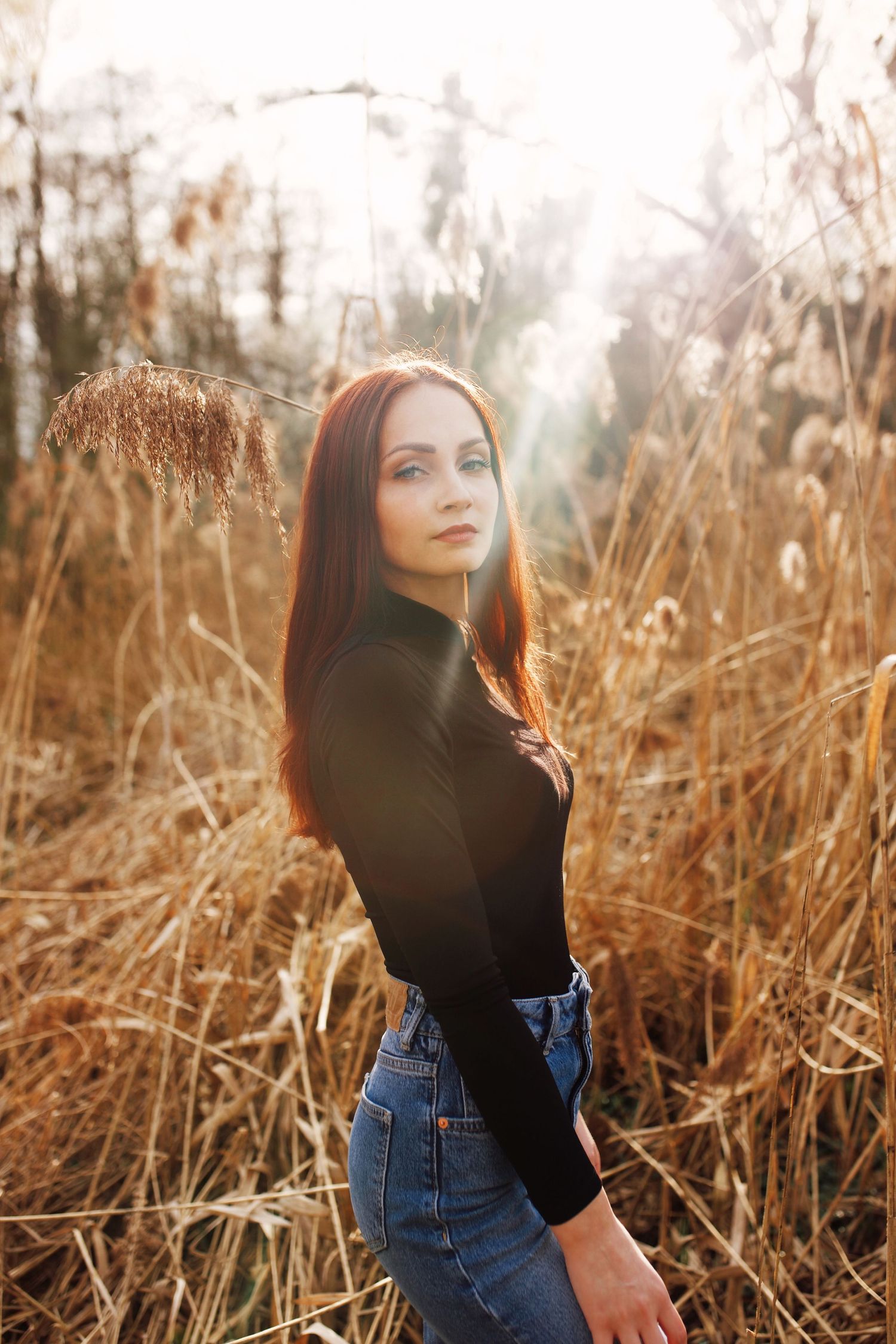 Young redhead woman in black turtleneck and jeans poses in golden dried grass field at sunset.
