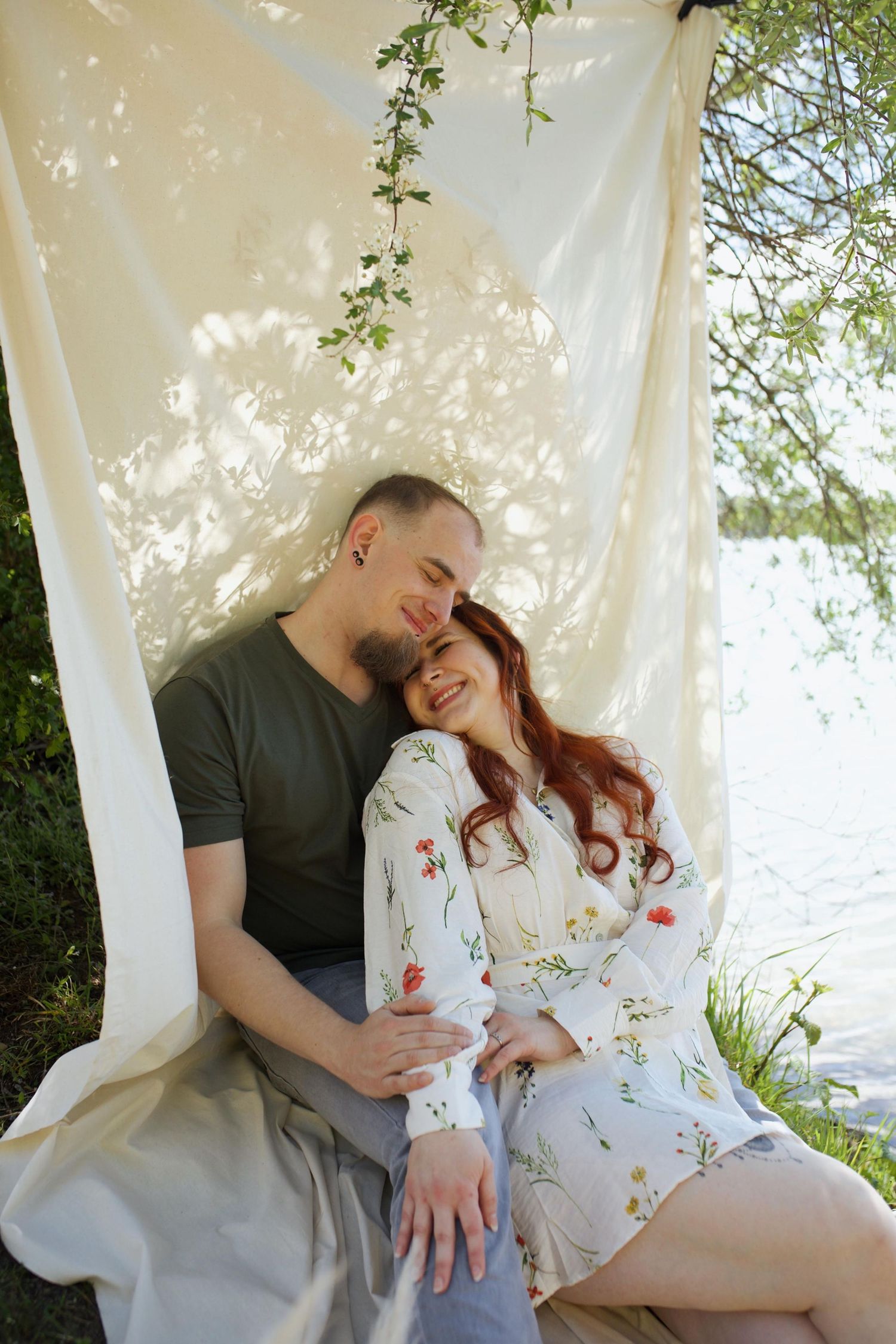 Romantic couple embracing under white canopy drapes near lake surrounded by lush green trees in sunlight.