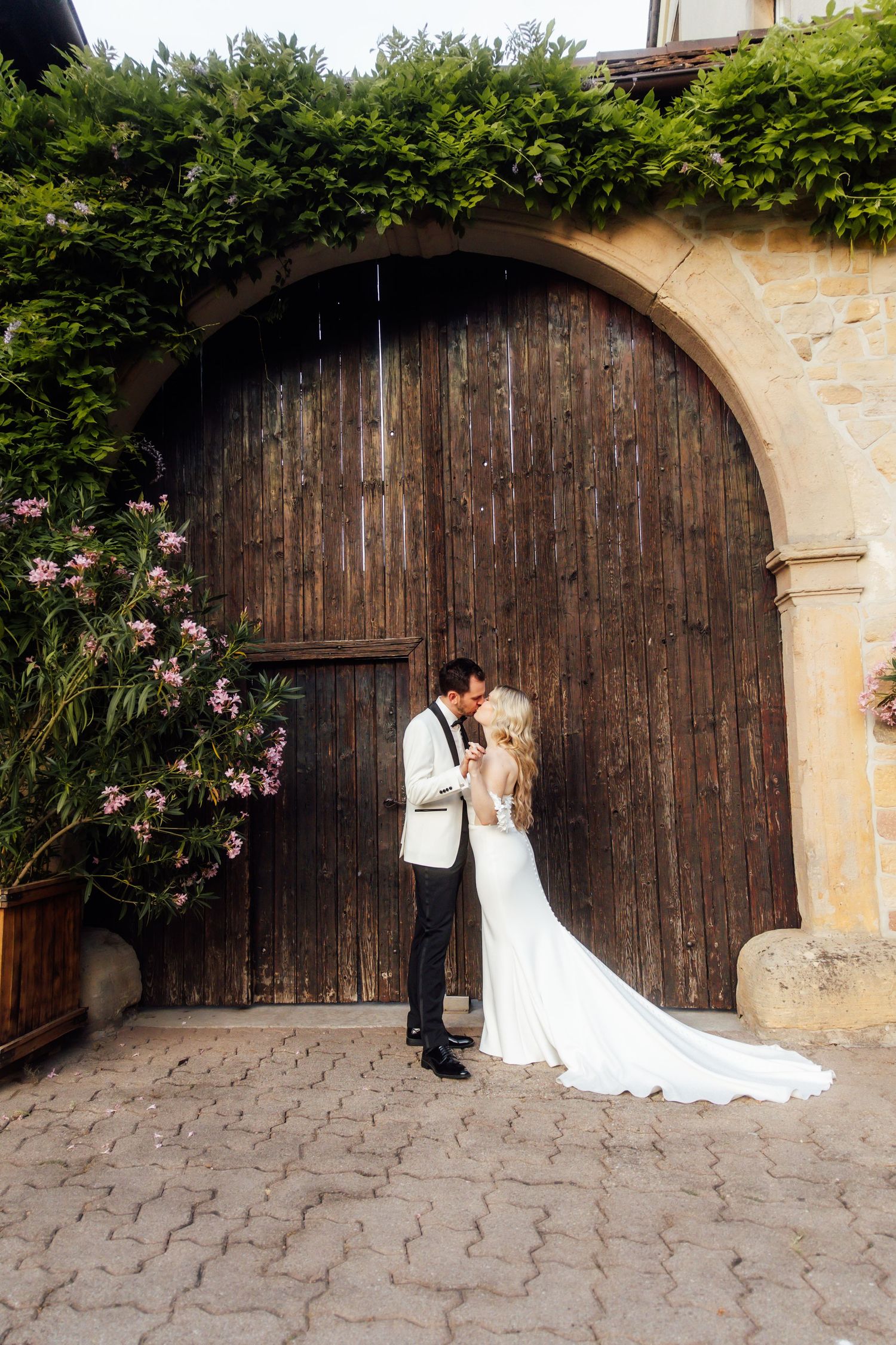 Bride and groom kissing in front of rustic wooden arched doors surrounded by lush greenery on cobblestone.
