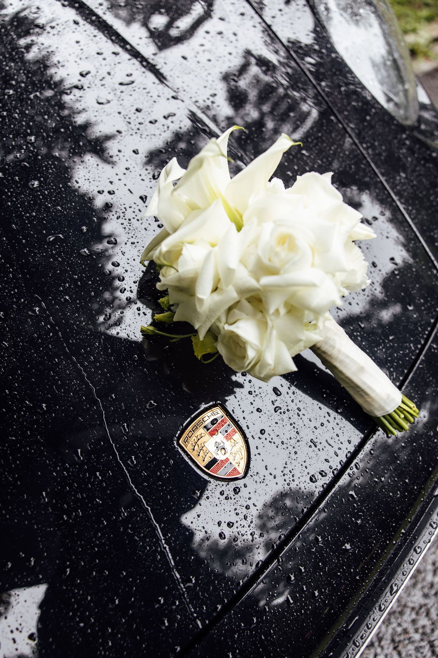 White wedding bouquet of roses and calla lilies resting on a wet black Porsche hood.