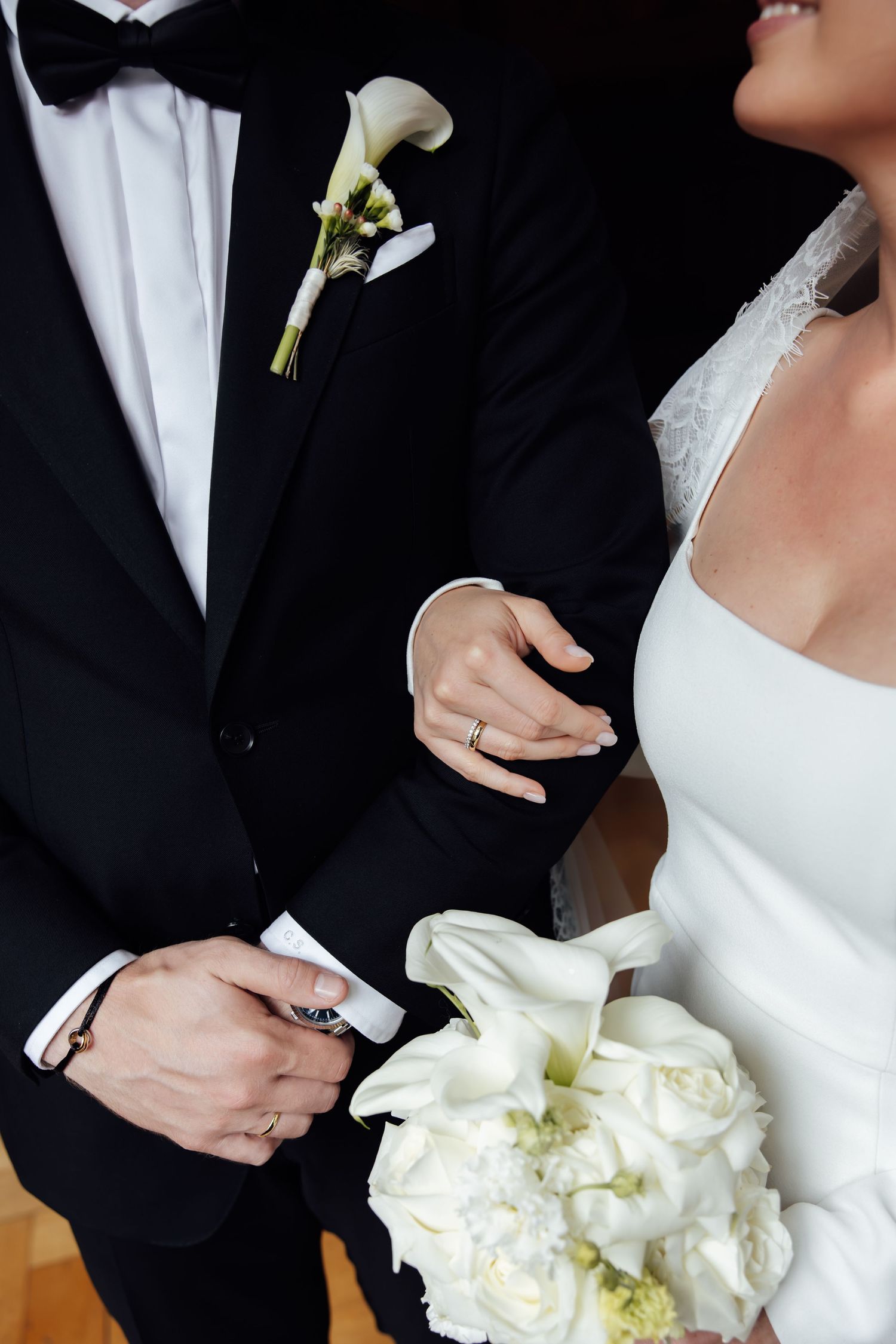 Bride and groom standing together, bride holding white calla lily bouquet, wearing wedding rings, groom in black tuxedo.