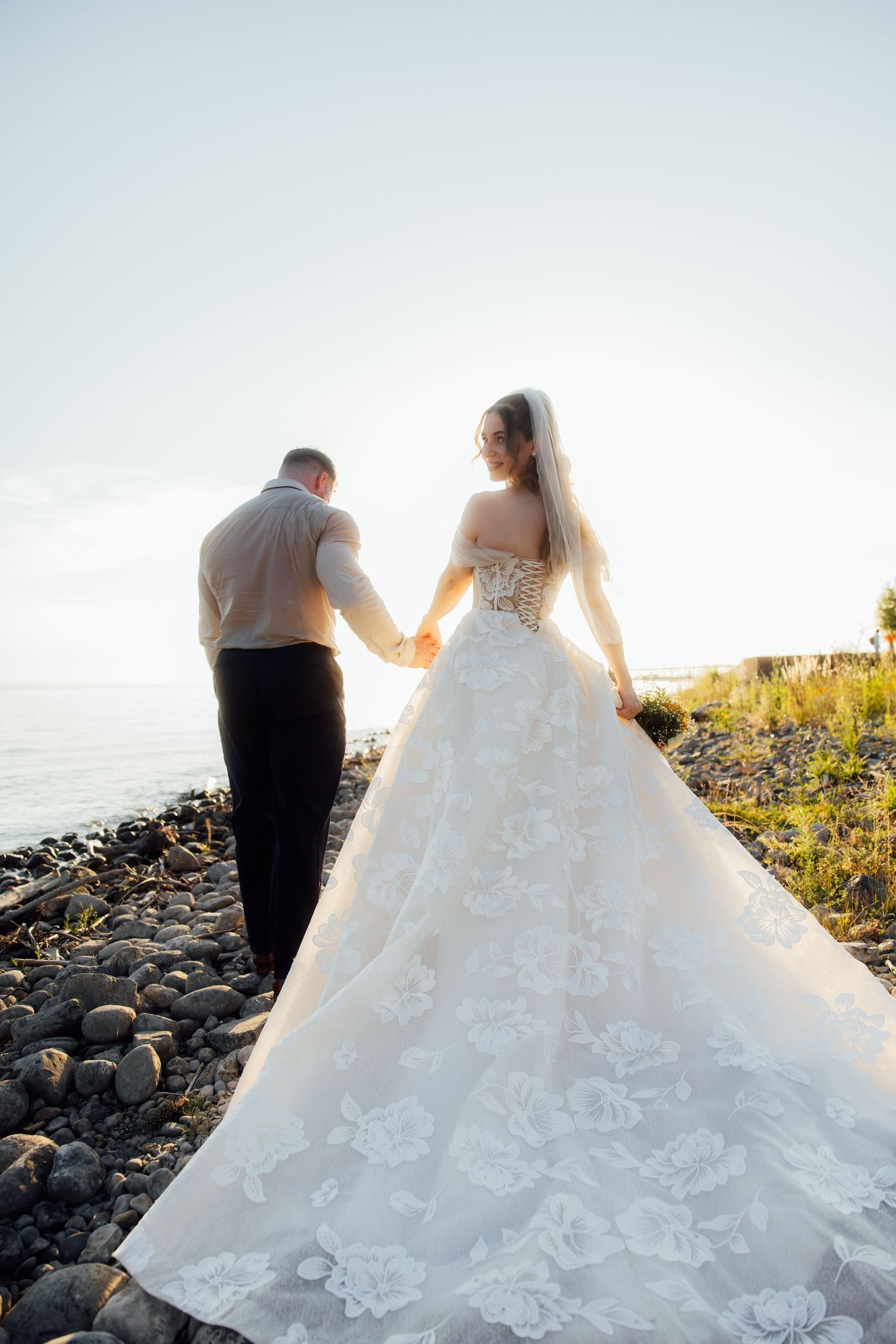 Bride and groom holding hands on rocky shore at sunset in floral wedding gown.