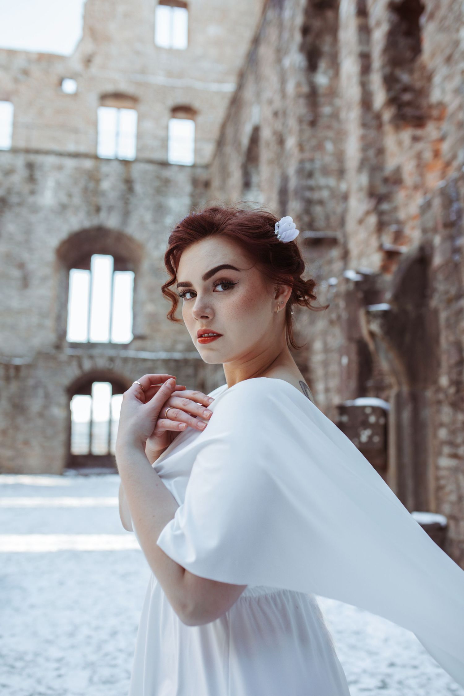 A woman in a flowing white dress with a flower in her hair poses in a historic stone ruin.