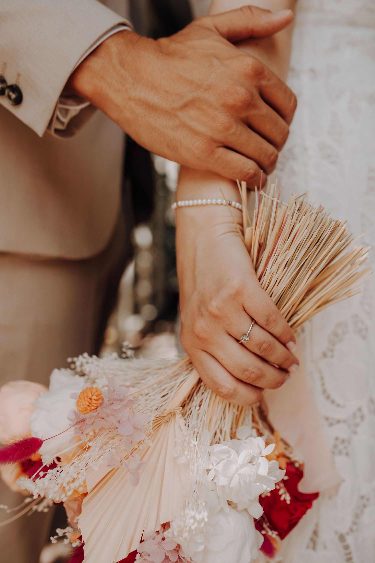 Close-up of bride and groom holding a dried floral bouquet, bride wearing a diamond ring and pearl bracelet.