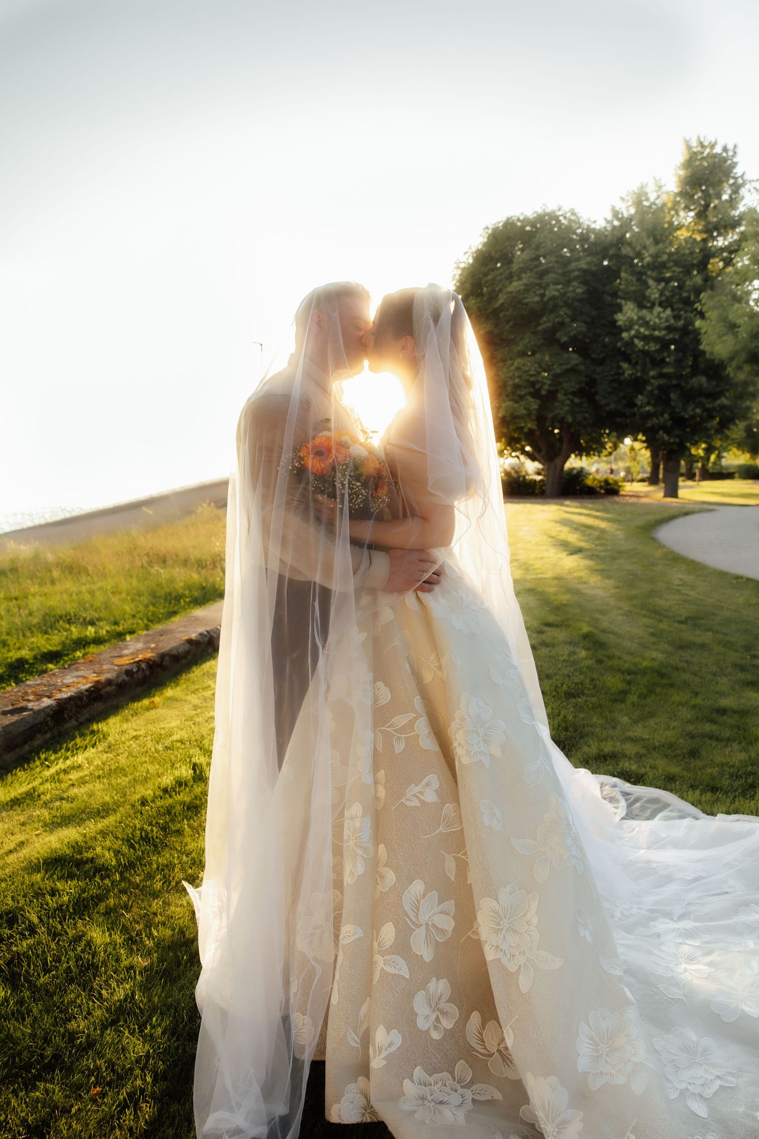 Bride and groom sharing a romantic kiss under a flowing veil with golden sunset backlight in a lush green outdoor setting.