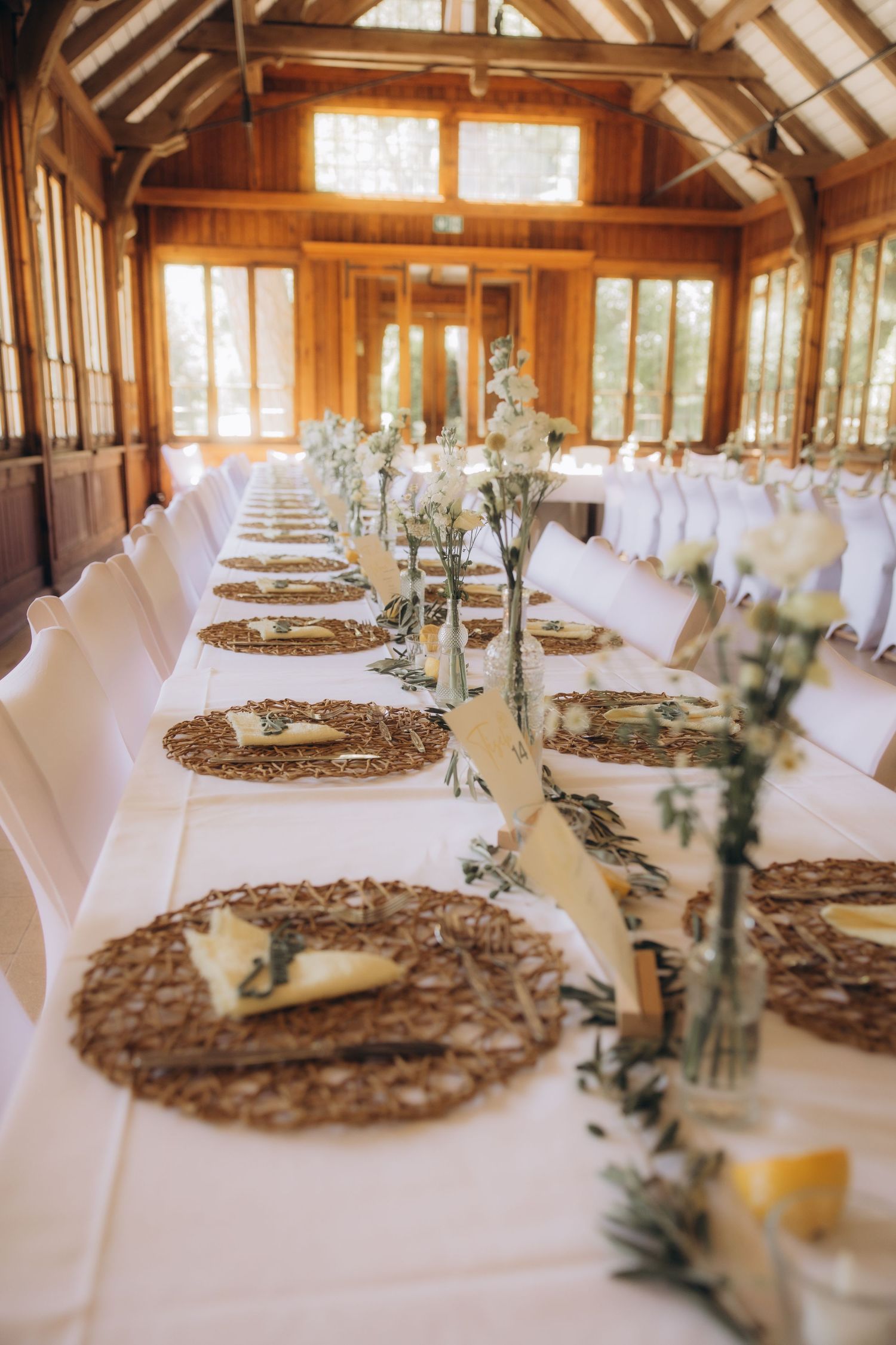 Elegant wedding reception table with wicker placemats, white chairs, floral centerpieces, and a white runner in a rustic wooden venue.