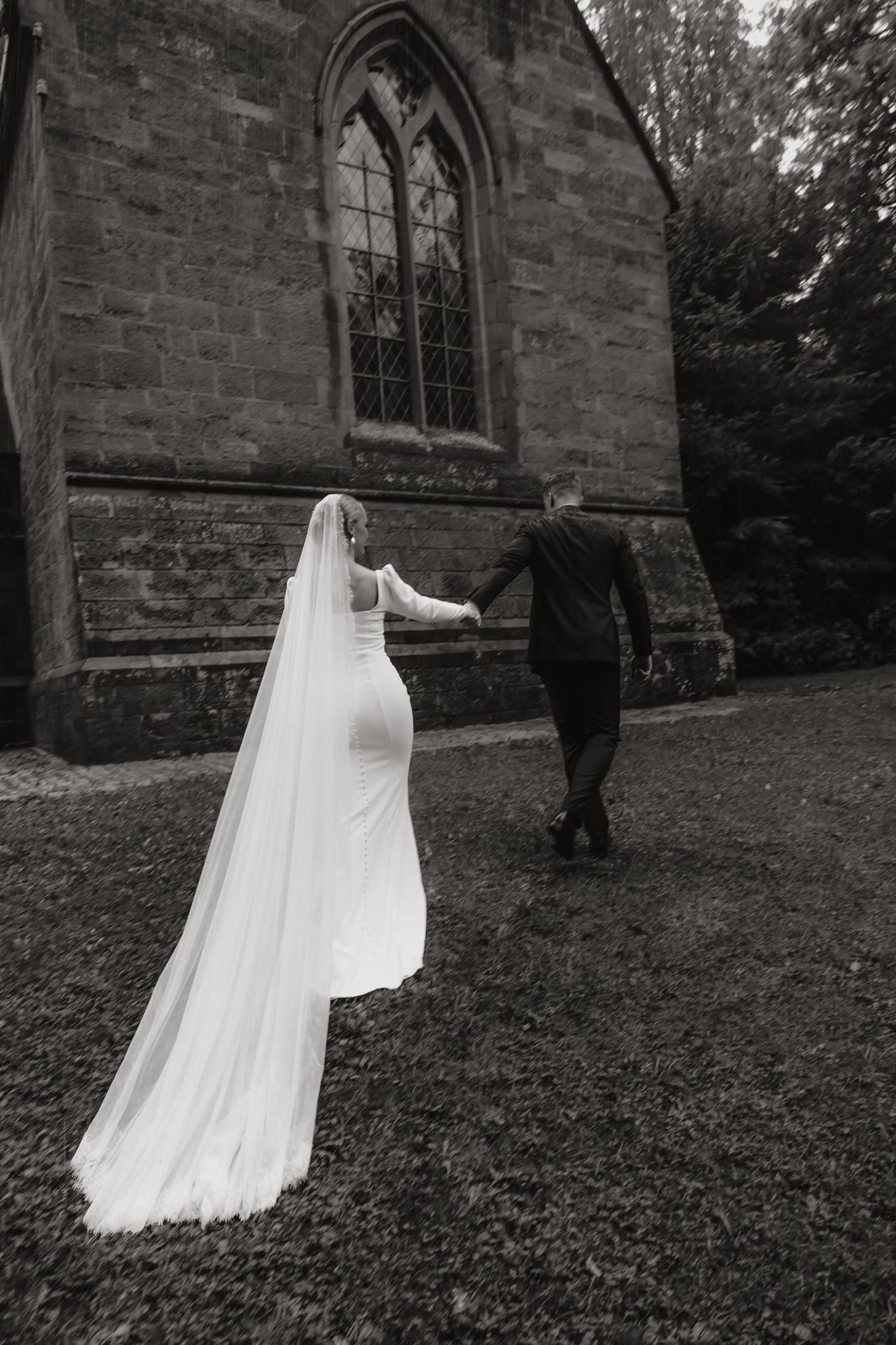 Black and white photo of a bride and groom holding hands walking toward a historic stone church.