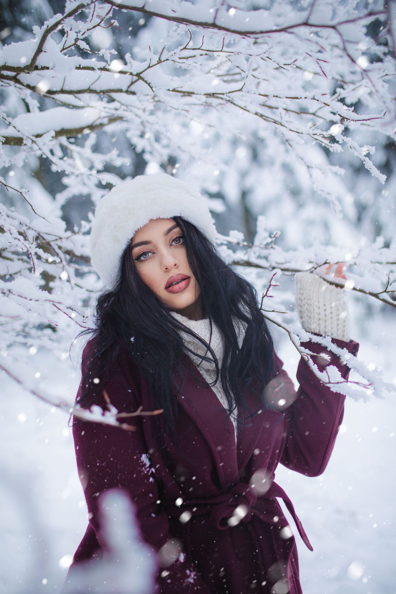 A dark-haired woman in a white fur hat and burgundy coat holds a cup in a snowy winter forest.