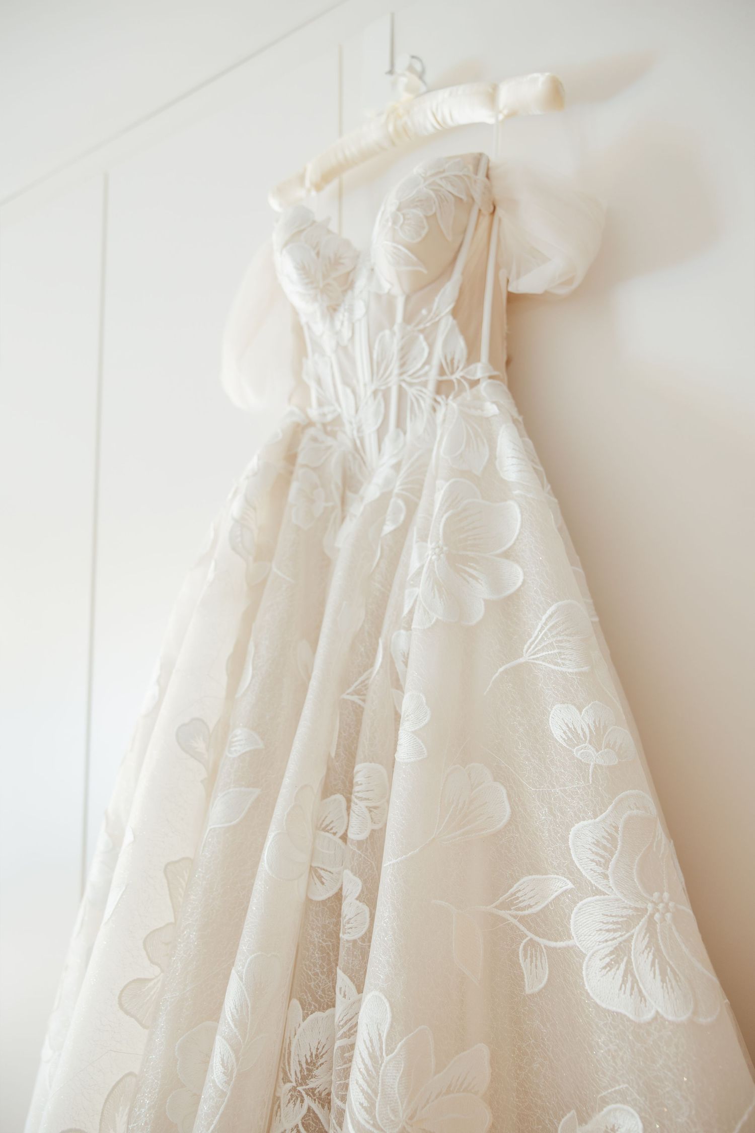 Elegant off-shoulder wedding gown with floral lace detail hanging on a white wall.