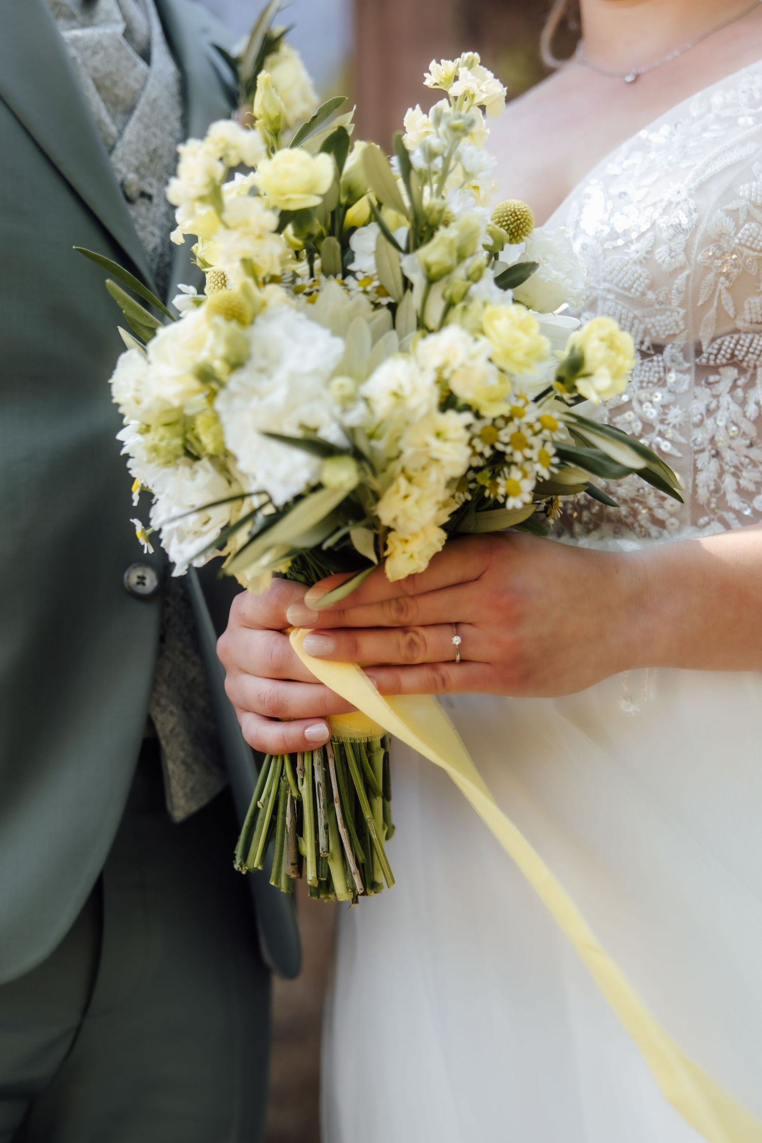 Bride and groom holding a white and yellow wedding bouquet with ribbon, bride wearing engagement ring and lace dress.