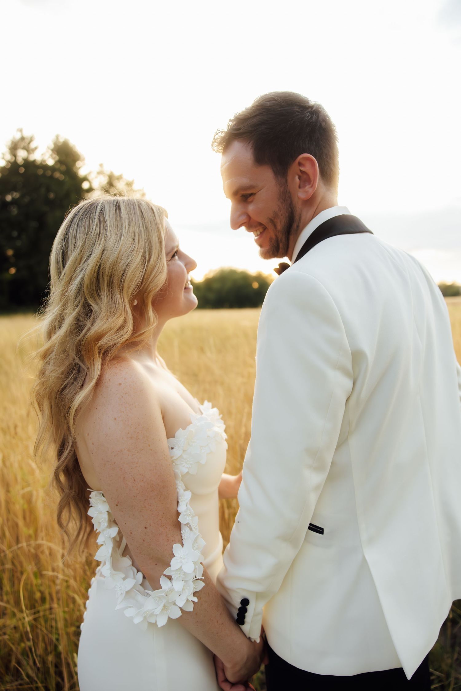 Bride and groom hold hands smiling at each other in golden field at sunset, both in white wedding attire.