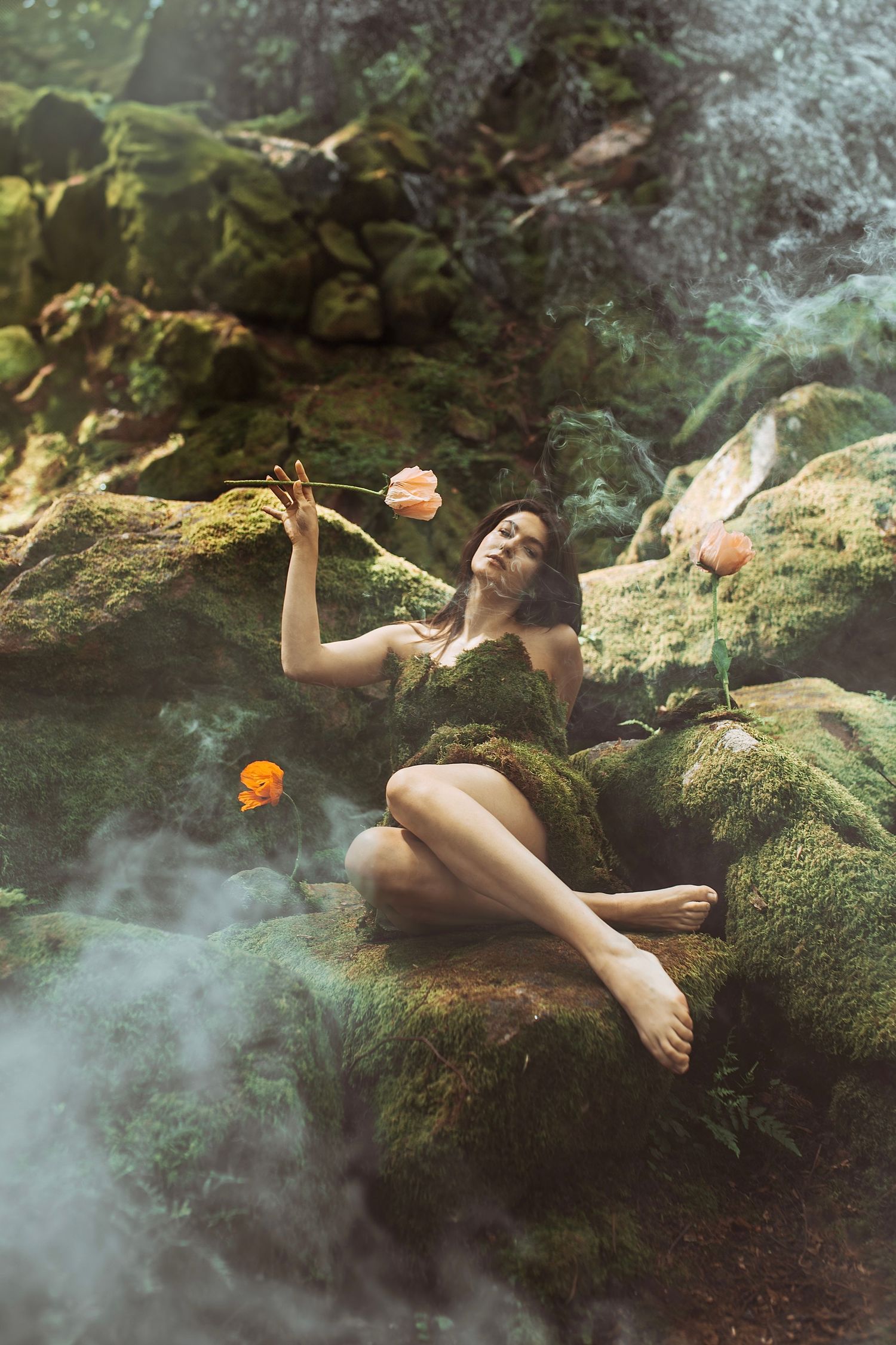 Woman in moss dress sits on mossy rocks holding orange tulips surrounded by mystical fog in enchanted forest.